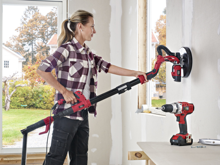 Eine Frau in kariertem Hemd und schwarzer Hose schleift eine Wand in einem Raum, der noch renoviert wird. In der Nähe befindet sich ein Tisch mit einer kabellosen Bohrmaschine und einigen Schleifscheiben. Durch das Fenster sind Bäume und Häuser zu sehen.