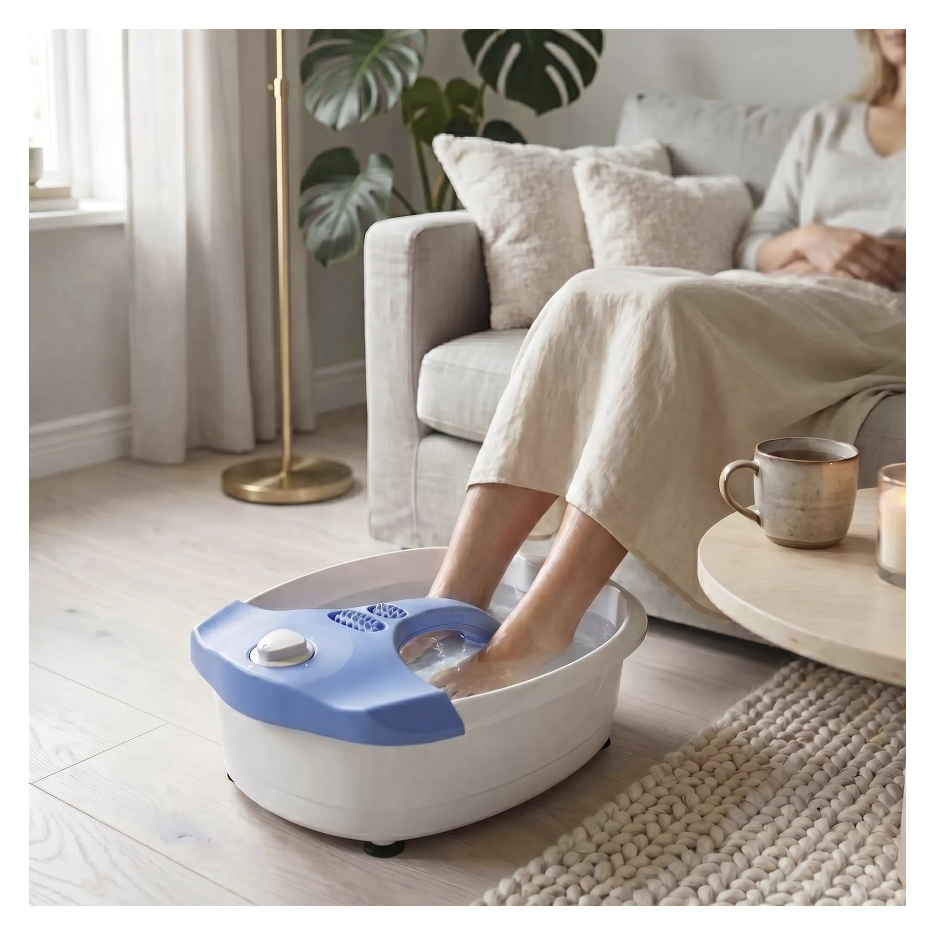 En person ligger i en soffa, med fötterna i Bright Foot-badet för massage. På soffbordet står en mugg, tallrik och tekanna. Det fridfulla rummet innehåller en matta, kuddar och en hög växt i bakgrunden.