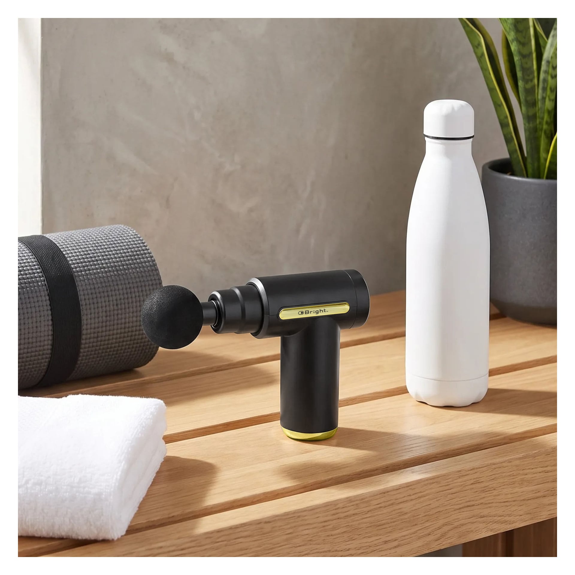 En svart Bright Massage Gun Mini med ett runt fäste står på en träbänk bredvid en grå yogamatta, en prydligt vikt vit handduk, en vit vattenflaska och en krukväxt i bakgrunden.