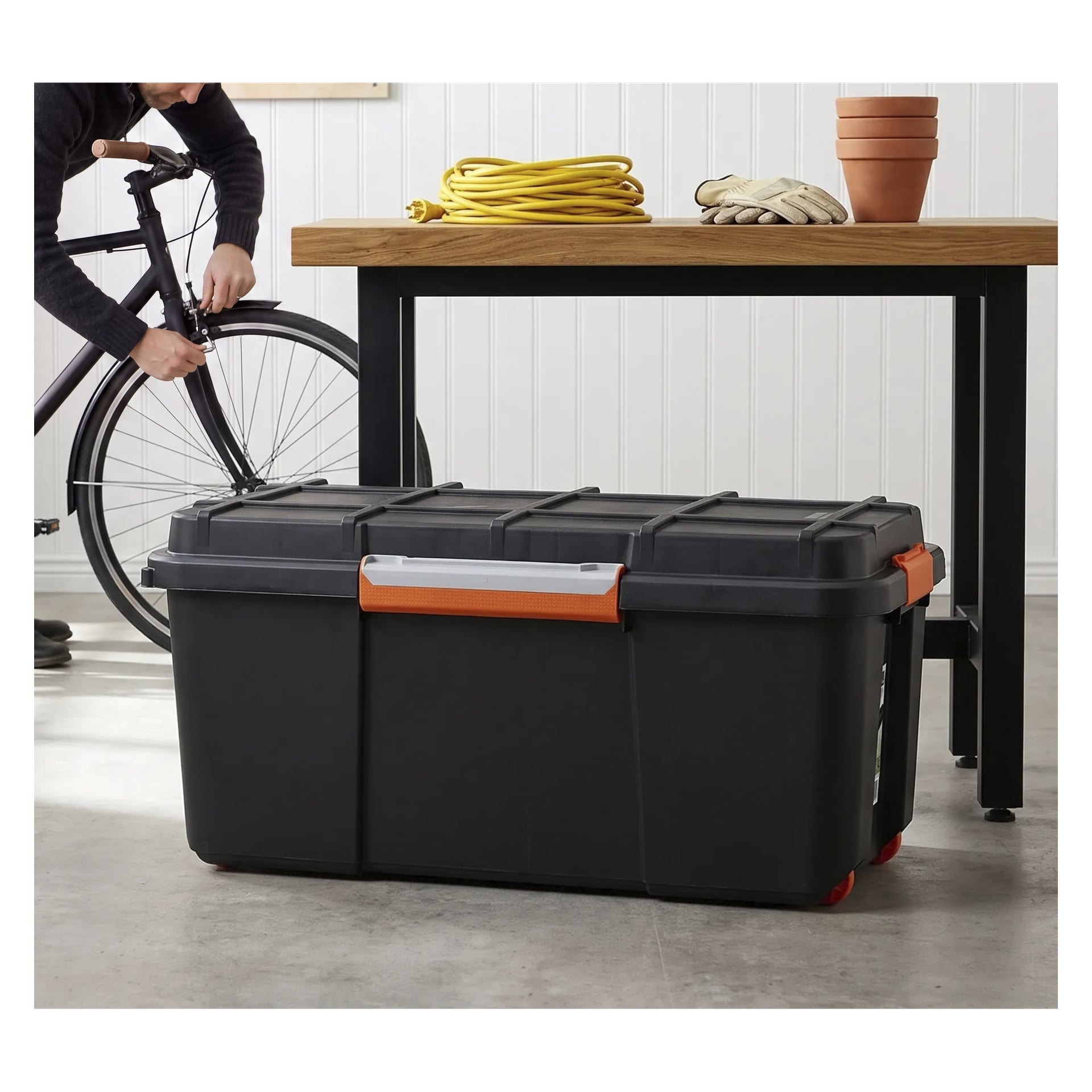 Joku työskentelee polkupyörän pyörän parissa Hard Head Storage -laatikon vieressä, jossa on musta muovirunko ja oranssit kahvat. Laatikko asetetaan lattialle puisen pöydän eteen, jossa on esineitä kuten keltainen köysi, hanskat ja useita pinottuja terrakottaruukkuja.
