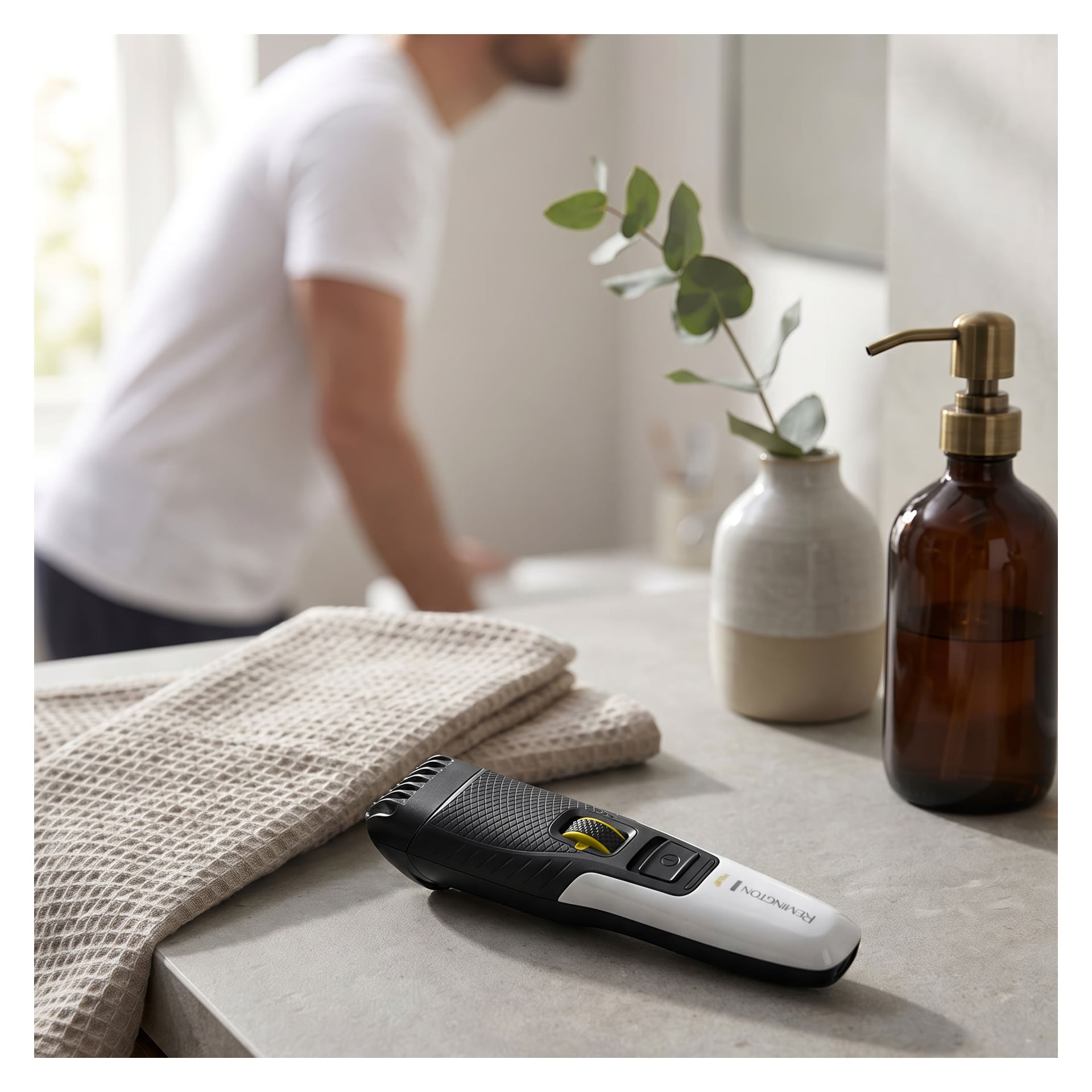 En svartvit Remington Cordless Beard Trimmer Style Series B4 står på en badrumsbänk bredvid en handduk, en brun tvålautomat och en vas fylld med gröna blad. En person i vit skjorta syns i den suddiga bakgrunden vid handfatet.