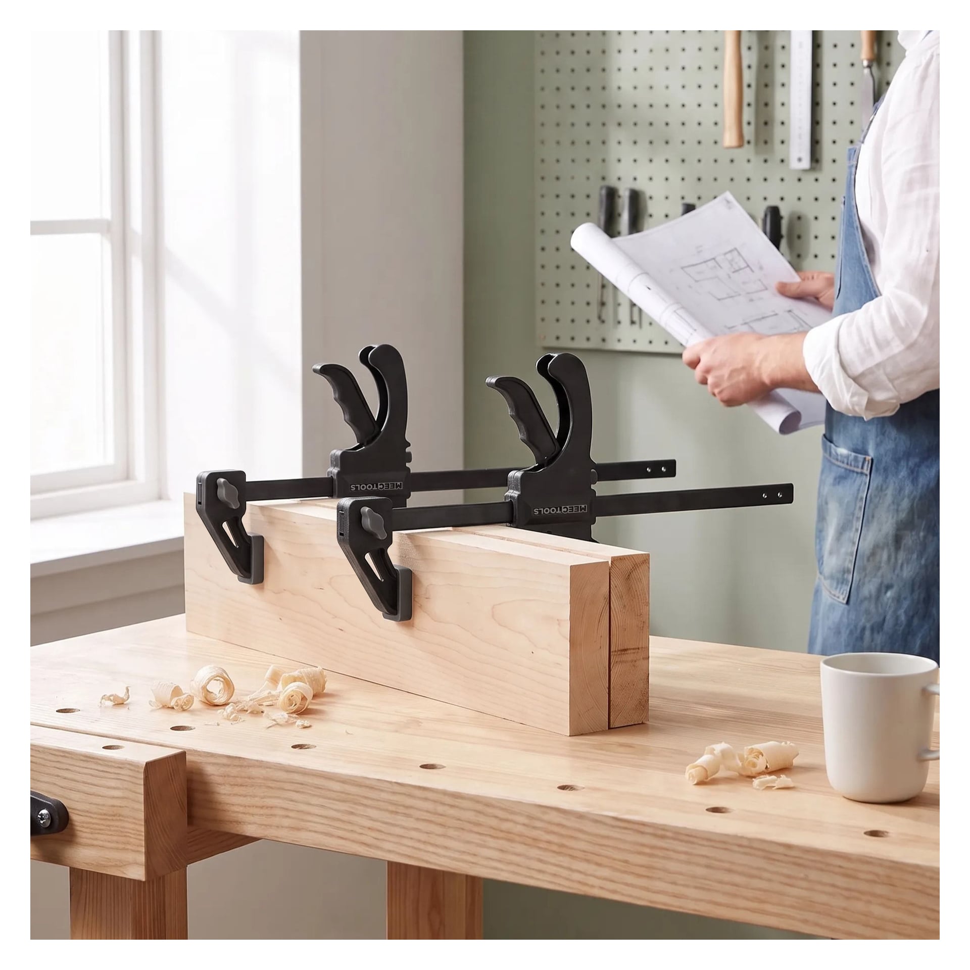 Två Meec Tools Quick Clamp 2-paket håller en träbalk på plats på en arbetsbänk. I bakgrunden finns träspån, en vit mugg och en person i förkläde som håller papper. Solljus från ett närliggande fönster lyser upp verktyg som hänger på en pegboard längs väggen.