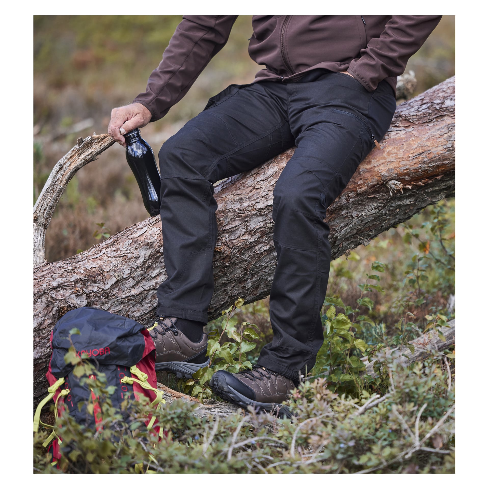 En person i "Kayoba Outdoor's Men Älvdalen 2.0"-bukser og brun jakke sitter på et veltet tre og holder en svart flaske. Tursko legges på bakken nær en rød og svart ryggsekk, alt omgitt av grønn utevegetasjon.