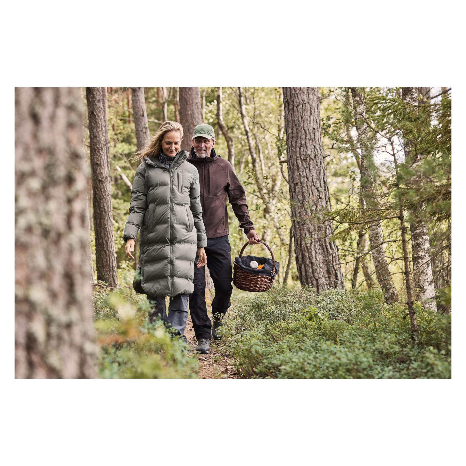 En kvinna i Kayoba Outdoor Women's winterrock Storlien promenerar längs en skogsstig, följd av en man i keps och jacka som bär en picknickkorg av rotting med saker inuti. Höga träd och tät grönska omger dem.