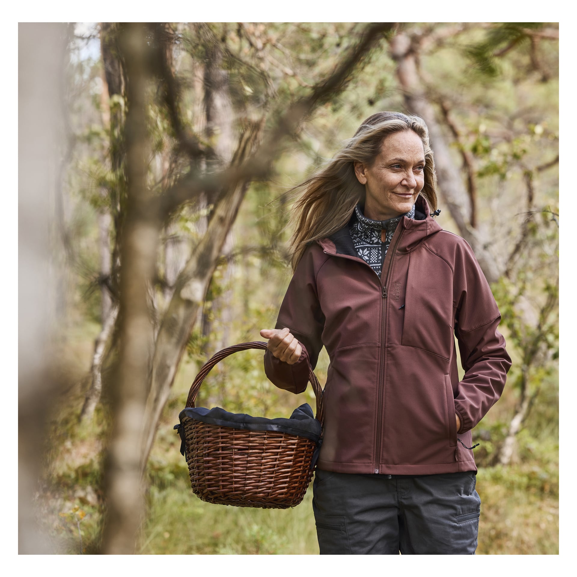 En kvinna i Kayoba Outdoor Women's fleecefodrade softshelljacka Hede står bland träd och grönska, med en korg av fläting i vänster hand. Hon tittar åt höger och ler svagt.