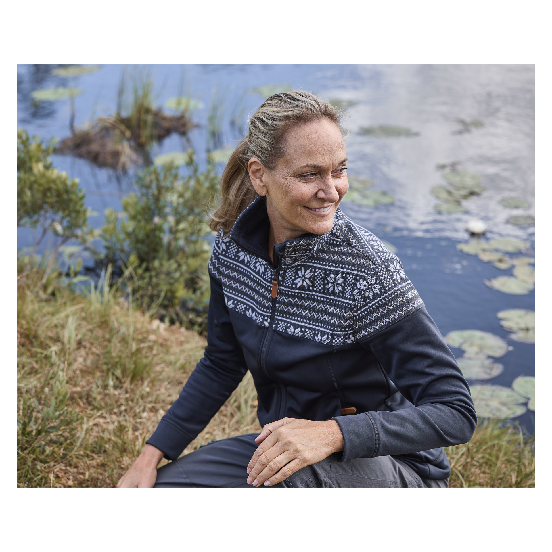 Eine lächelnde Frau mit blondem Haar, das zu einem Pferdeschwanz gebunden ist, sitzt auf dem Gras neben einem mit Seerosen übersäten Teich, gekleidet in den Kayoba Outdoor Women's Valbo Mid-Layer-Pullover, der einen Reißverschluss vorne und weiße nordische Muster hat. Sie blickt zur Seite, umgeben von üppigem Grün und reflektierendem Wasser.