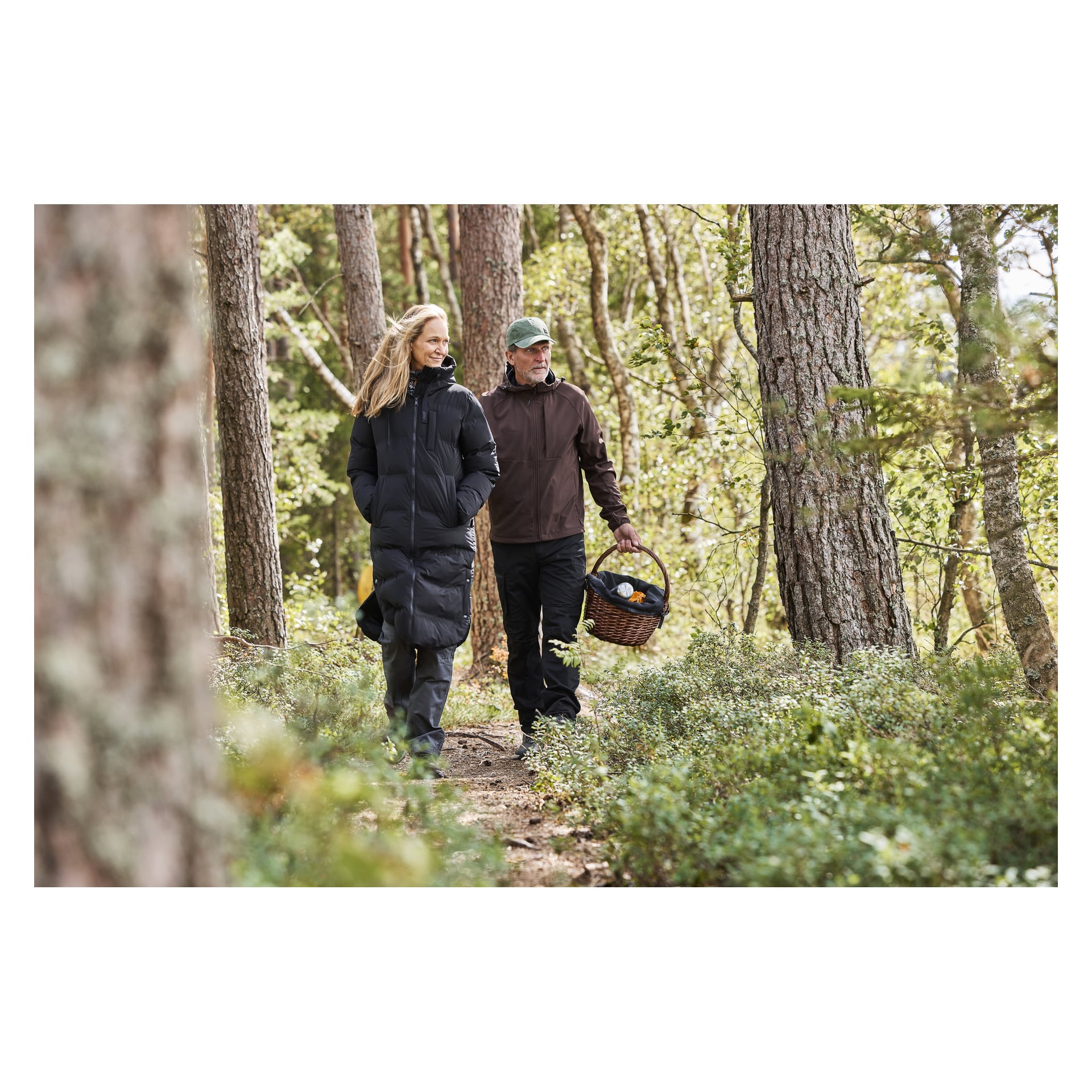 En man och en kvinna promenerar genom en skog omgivna av träd och frodig grönska. Kvinnan är klädd i Kayoba Outdoor Women's vinterrock Storlien, medan mannen bär en brun jacka, grön mössa och bär en korg av rotting.