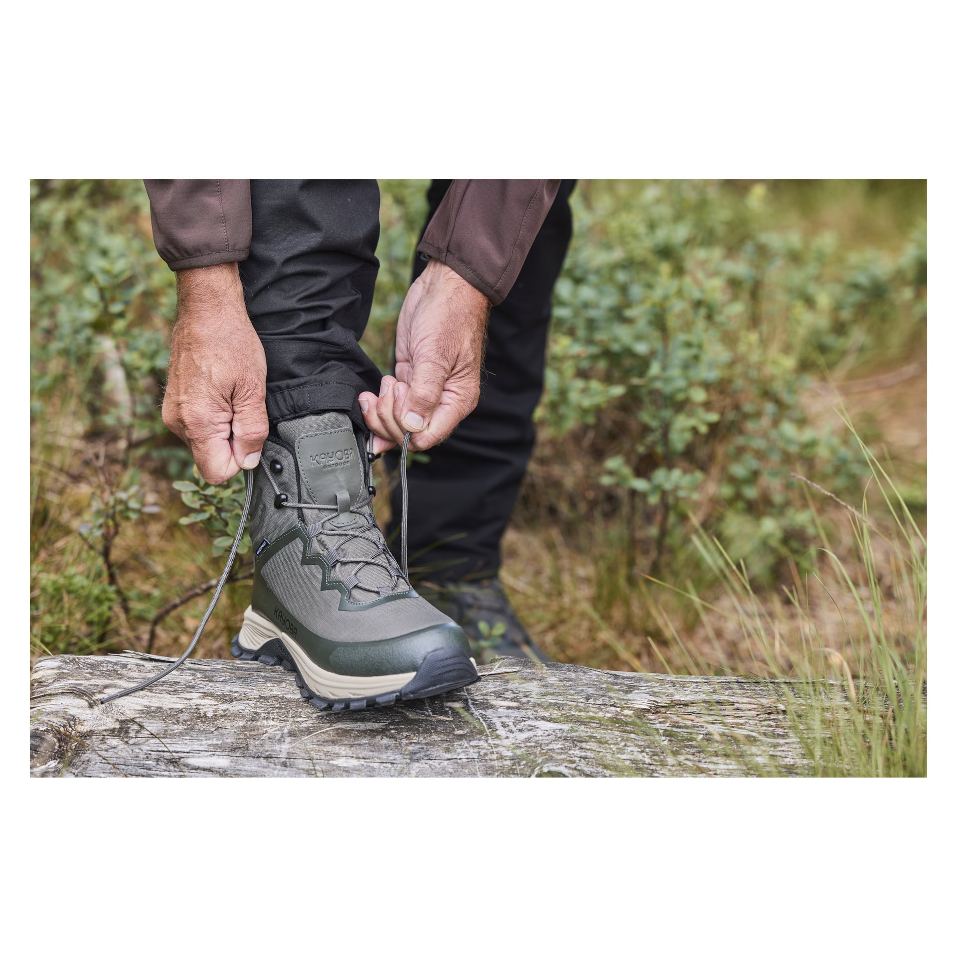 Iklädd svarta byxor och brun jacka knyter någon snörena på Kayoba Outdoor Waterproof vandringskängor medan de står på en nedfallen stock bland frodigt grönt växtlighet. Endast deras händer kan ses när de gör sig redo för ett utomhusäventyr.