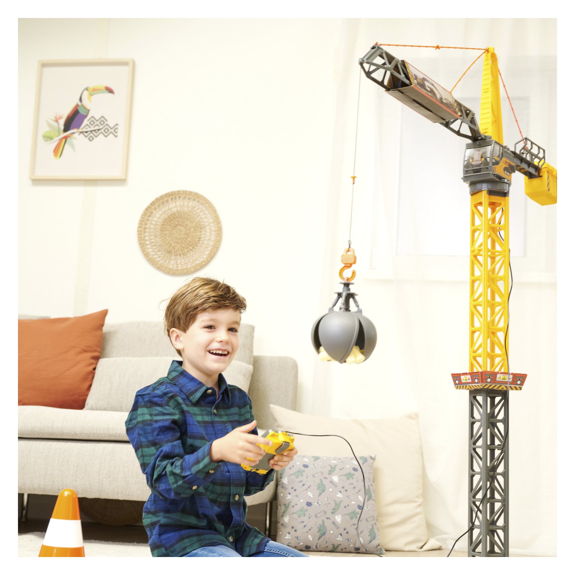 En glad ung pojke sitter på golvet inne och leker med Dickie Toys Mighty Crane, en distansstyrd byggleksak. Bakom honom finns en soffa, en kudde, några väggdekorationer och en liten trafikkon.