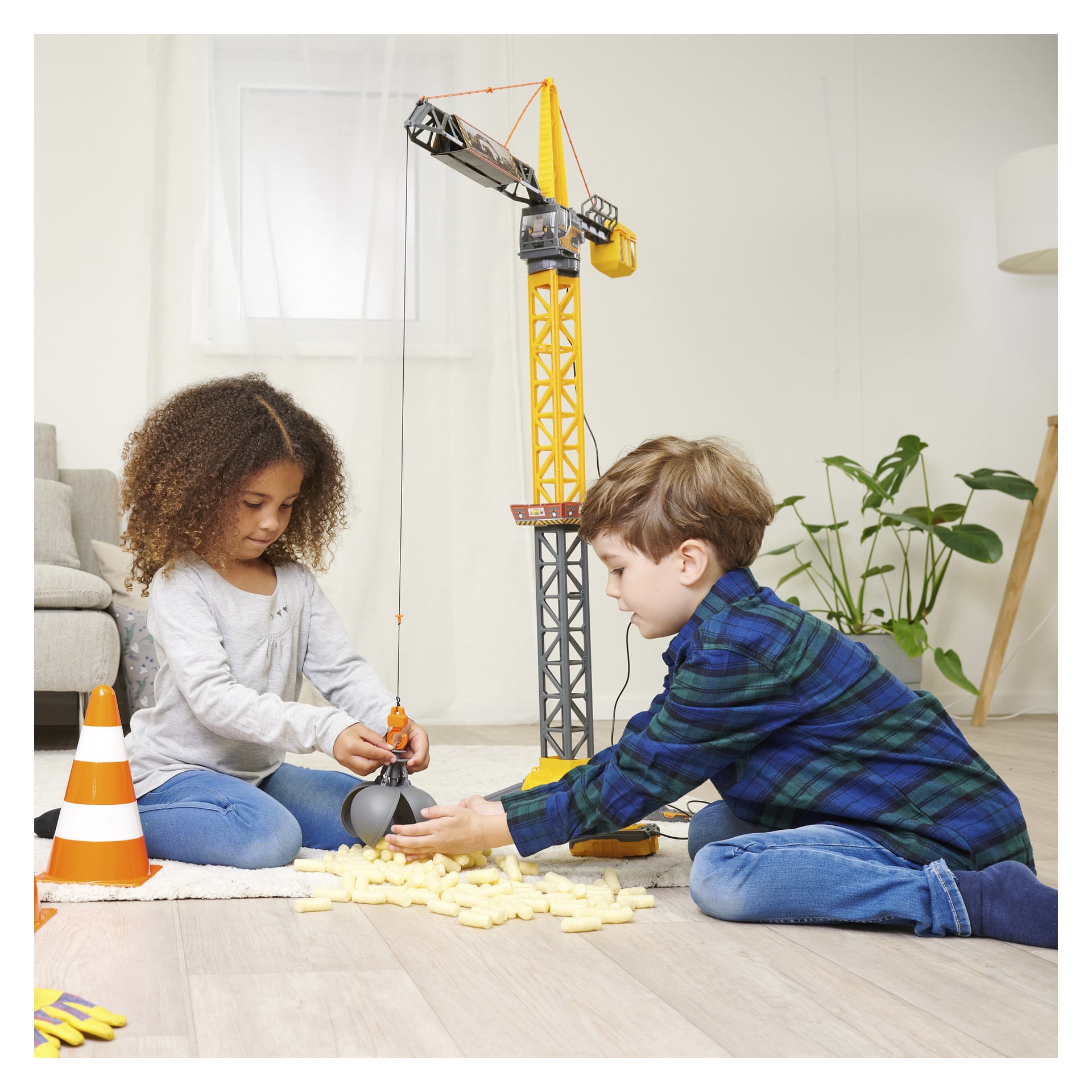 En flicka och en pojke sitter på golvet och leker med en Dickie Toys radiostyrd Mighty Crane och skumblock. Flickan justerar kranens krok medan pojken staplar block i närheten. I bakgrunden finns en trafikkon och en krukväxt.