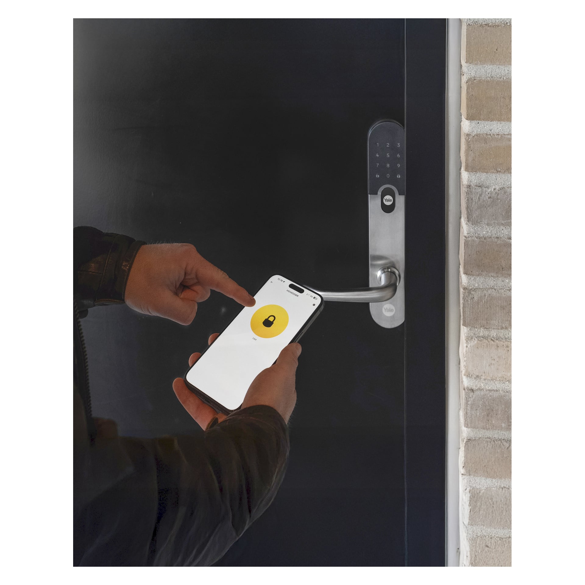 En person står foran en svart dør med en Yale Entrus digital lås, og holder en smarttelefon som viser en app med et gult låseikon.