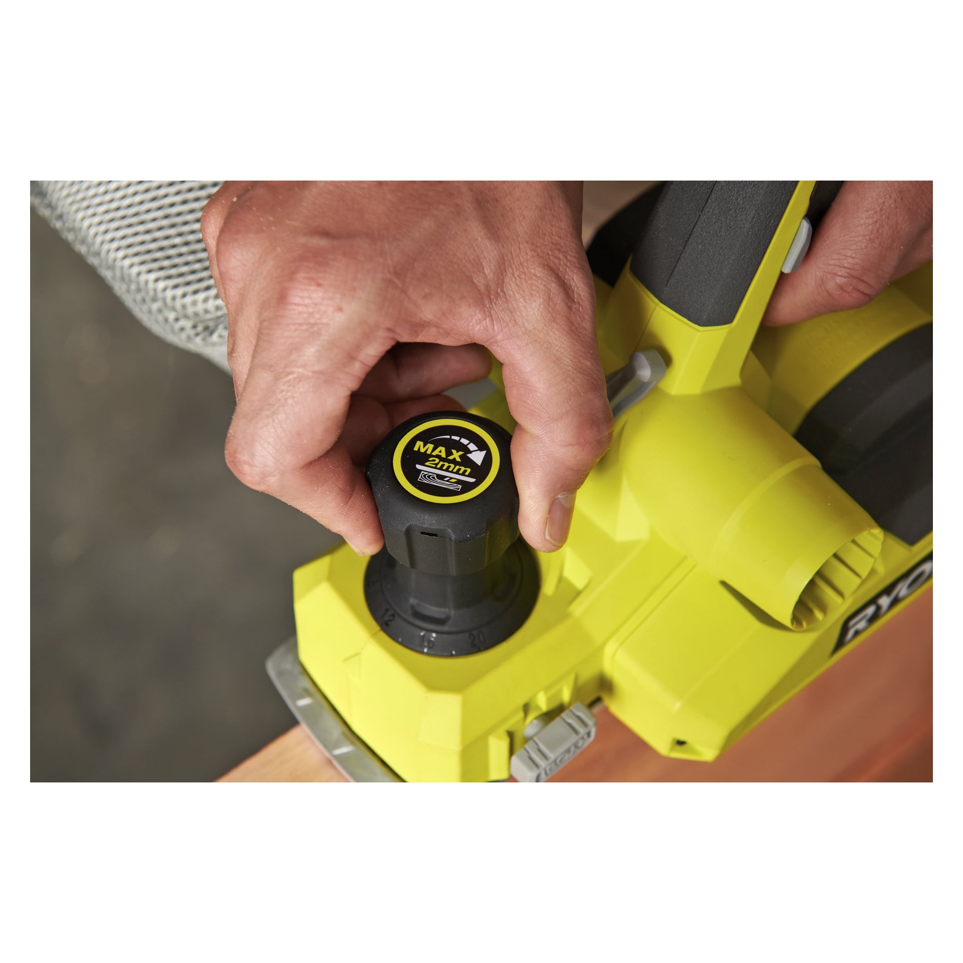 Ktoś przekręca pokrętło kontroli głębokości w żółtej strugarce elektrycznej Ryobi One+ Cordless RPL18-0, ustawiając ją na 2 mm podczas strugania kawałka drewna.