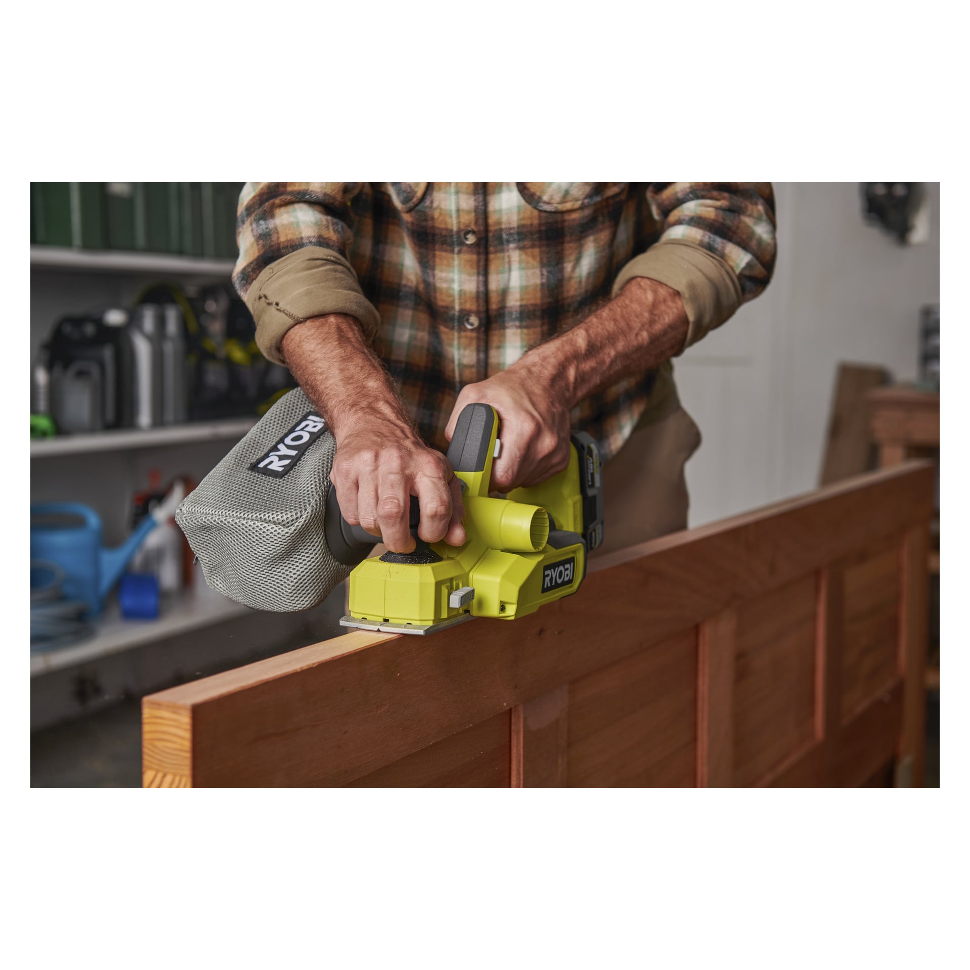 Ubrana w koszulę w kratę, osoba używa Ryobi One+ Cordless Electric Planer RPL18-0, aby strugować krawędź drewnianych drzwi w warsztacie, pokazując jedynie tułów i dłonie.