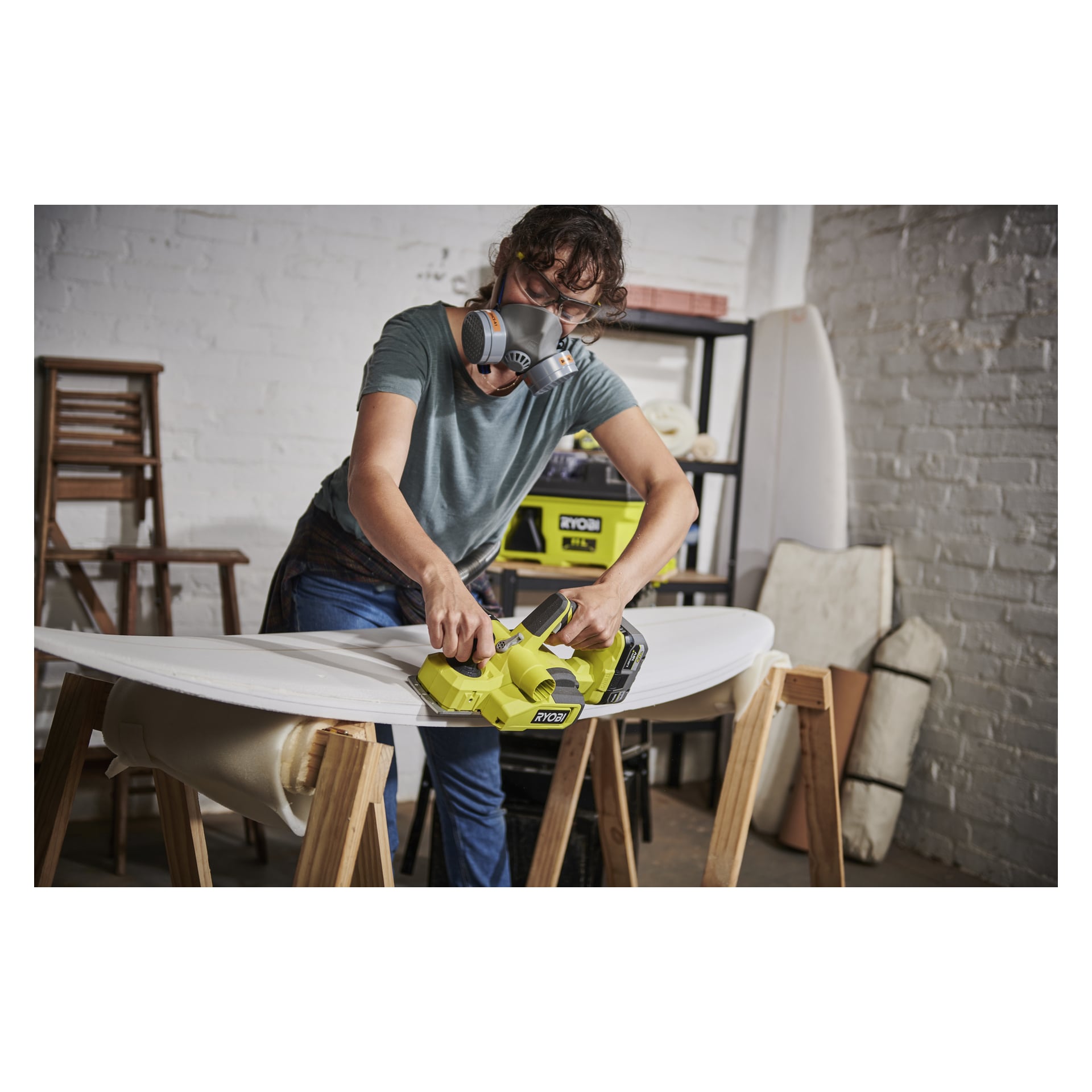 Osoba nosząca maskę respiratora kształtuje lub szlifuje deskę surfingową podtrzymywaną przez dwa drewniane stojaki, używając Ryobi One+ Cordless Electric Planer RPL18-0 w warsztacie z białymi ceglanymi ścianami.