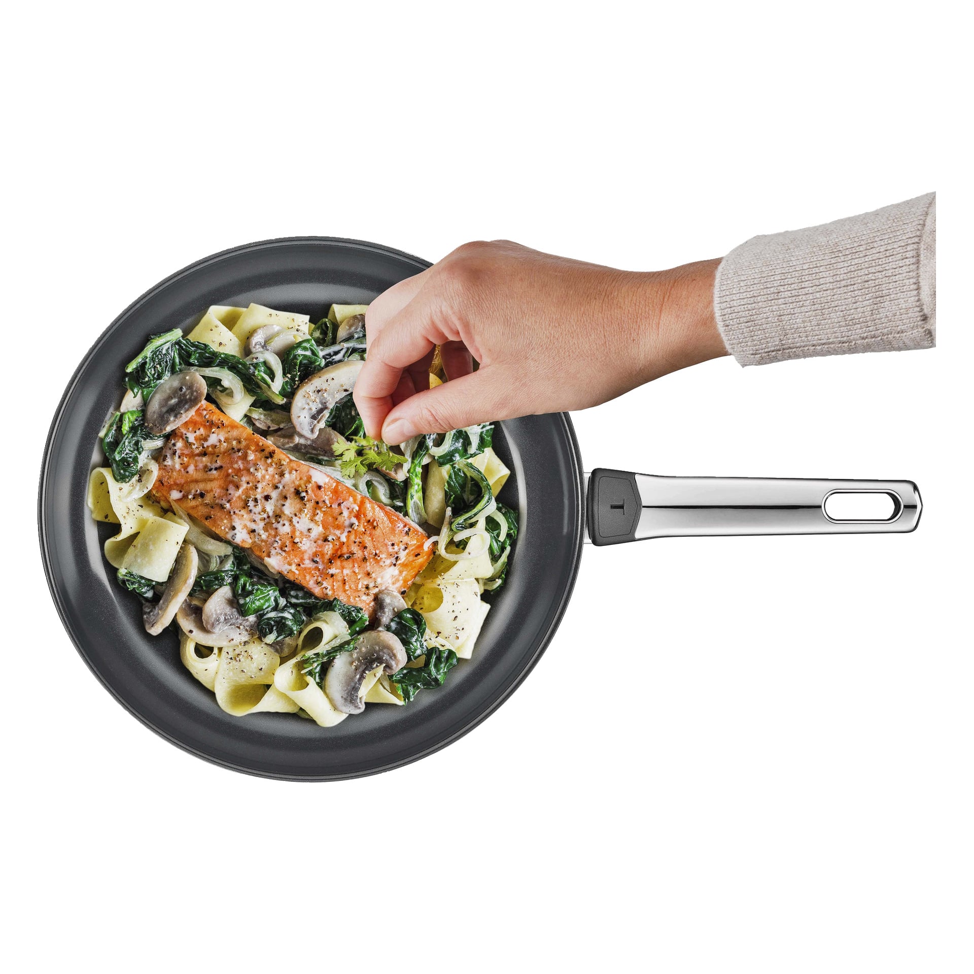 En hand tillsätter örter till lax med pasta, spenat och svamp i en Tefal stekpanna Emotion Plus SS.