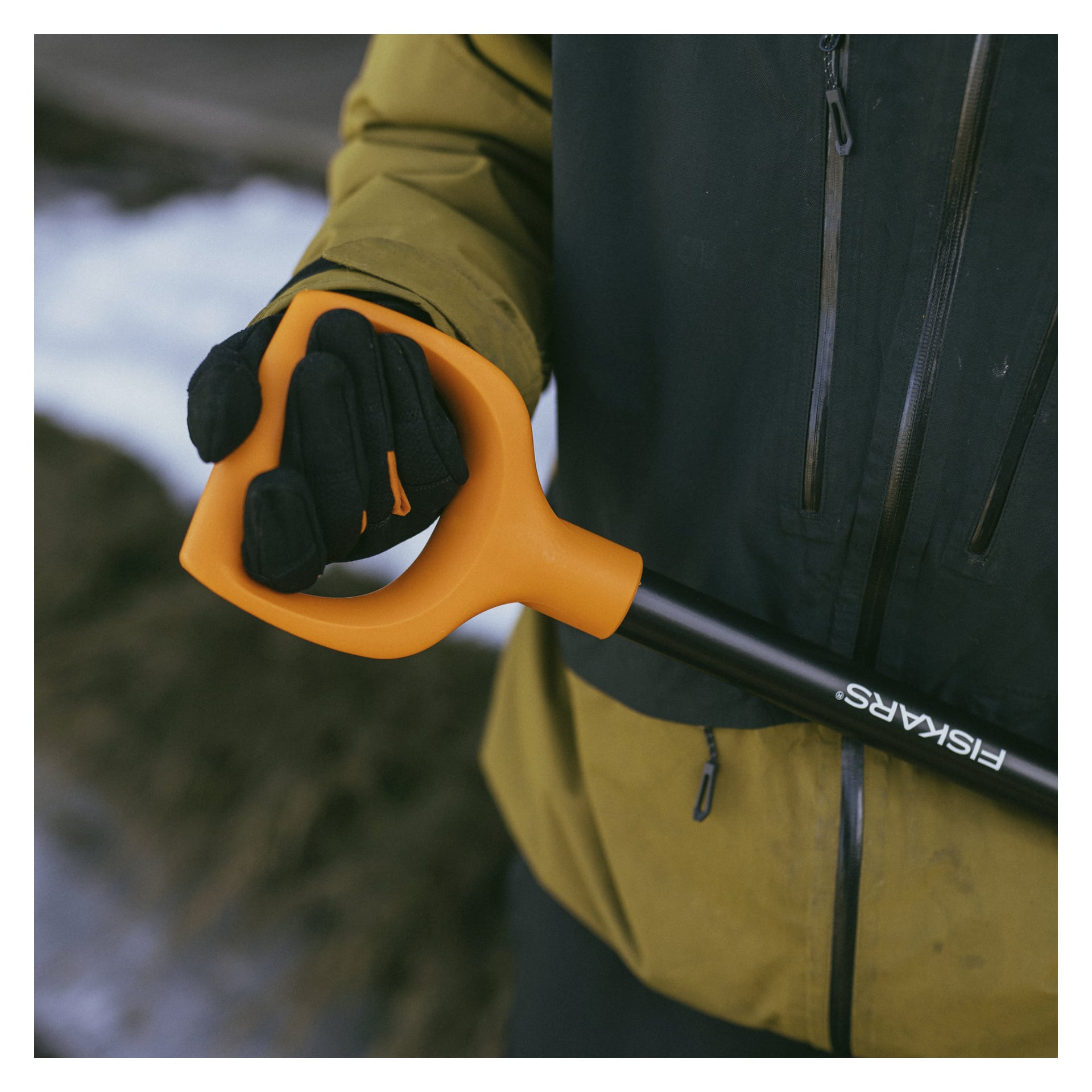 Någon i grön jacka och mörk handske håller i det orangea handtaget på en Fiskars is- och barkskrapa, med snö och jord suddiga i bakgrunden.