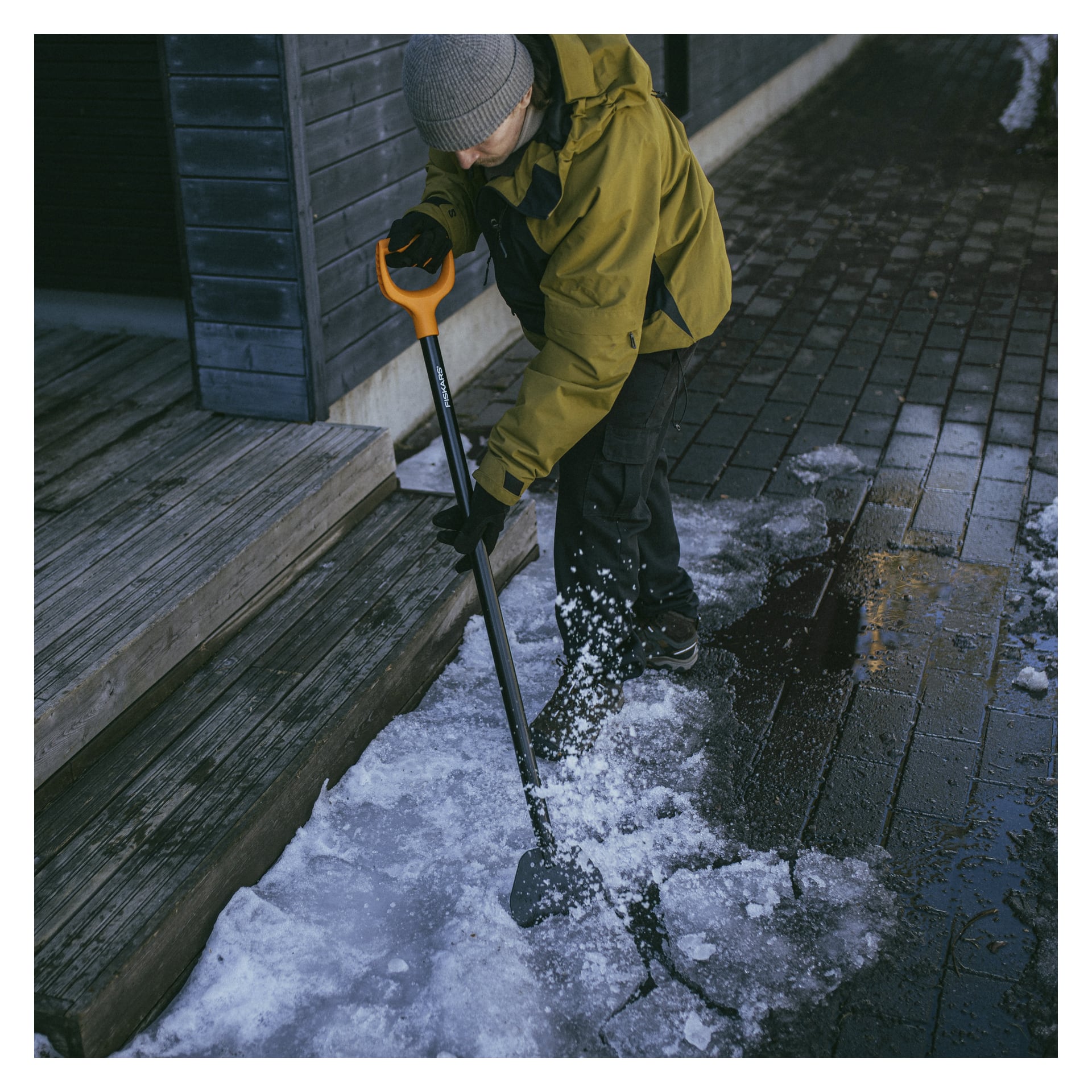 En person klädd i en grön jacka, svarta byxor och en grå mössa använder en Fiskars Ice and Bark Scraper för att hacka bort is på en tegelgångväg bredvid trätrappor. Området runt dem är fuktigt med fläckar av kvarvarande is.