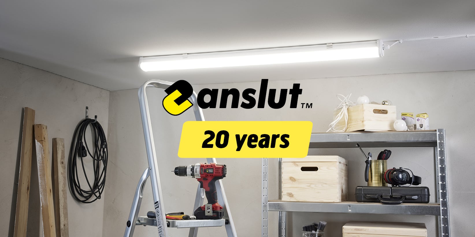 A garage or workshop featuring a metal ladder, a red drill, shelves stacked with wooden boxes, various tools and bulbs, and a fluorescent light overhead. The words “anslut 20 years” appear in black and yellow.