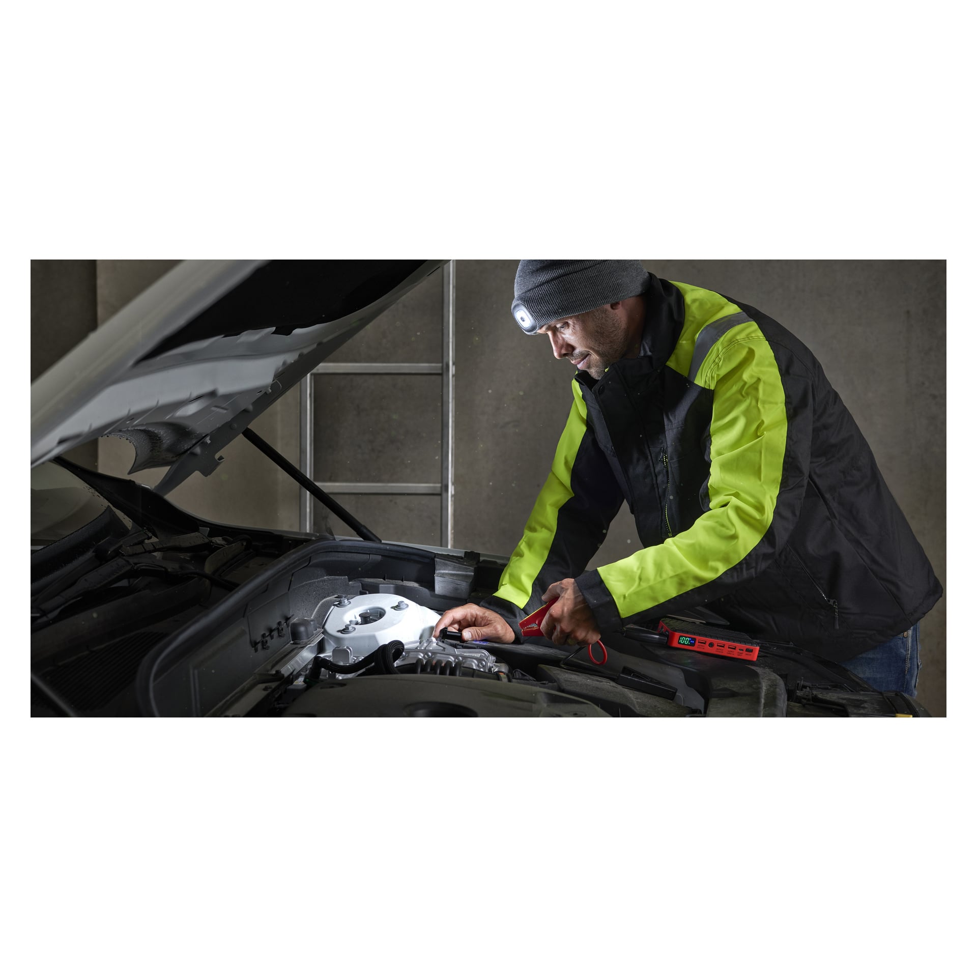 Ein Mann mit Stirnlampe benutzt einen Hamron-Jumpstarter mit Powerbank, um Kabel mit einer Autobatterie unter einer offenen Motorhaube zu verbinden.