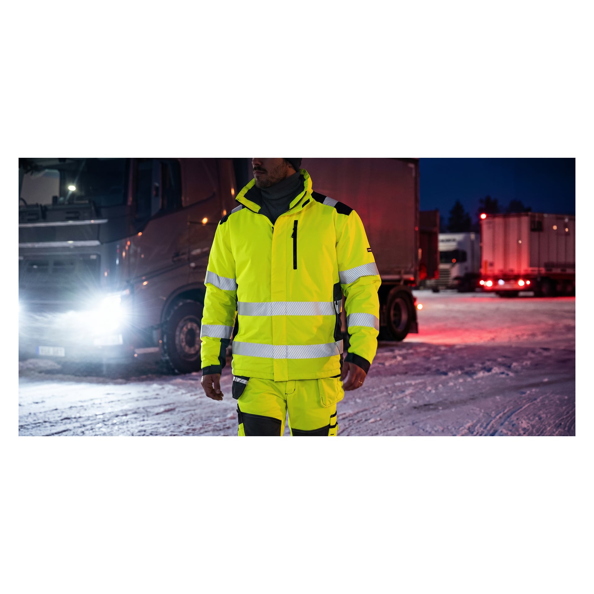 En person som bär en Bluewear Warm-fodrad Hi-Vis-jacka med reflekterande ränder och matchande byxor står framför parkerade lastbilar på en snötäckt yta på natten.