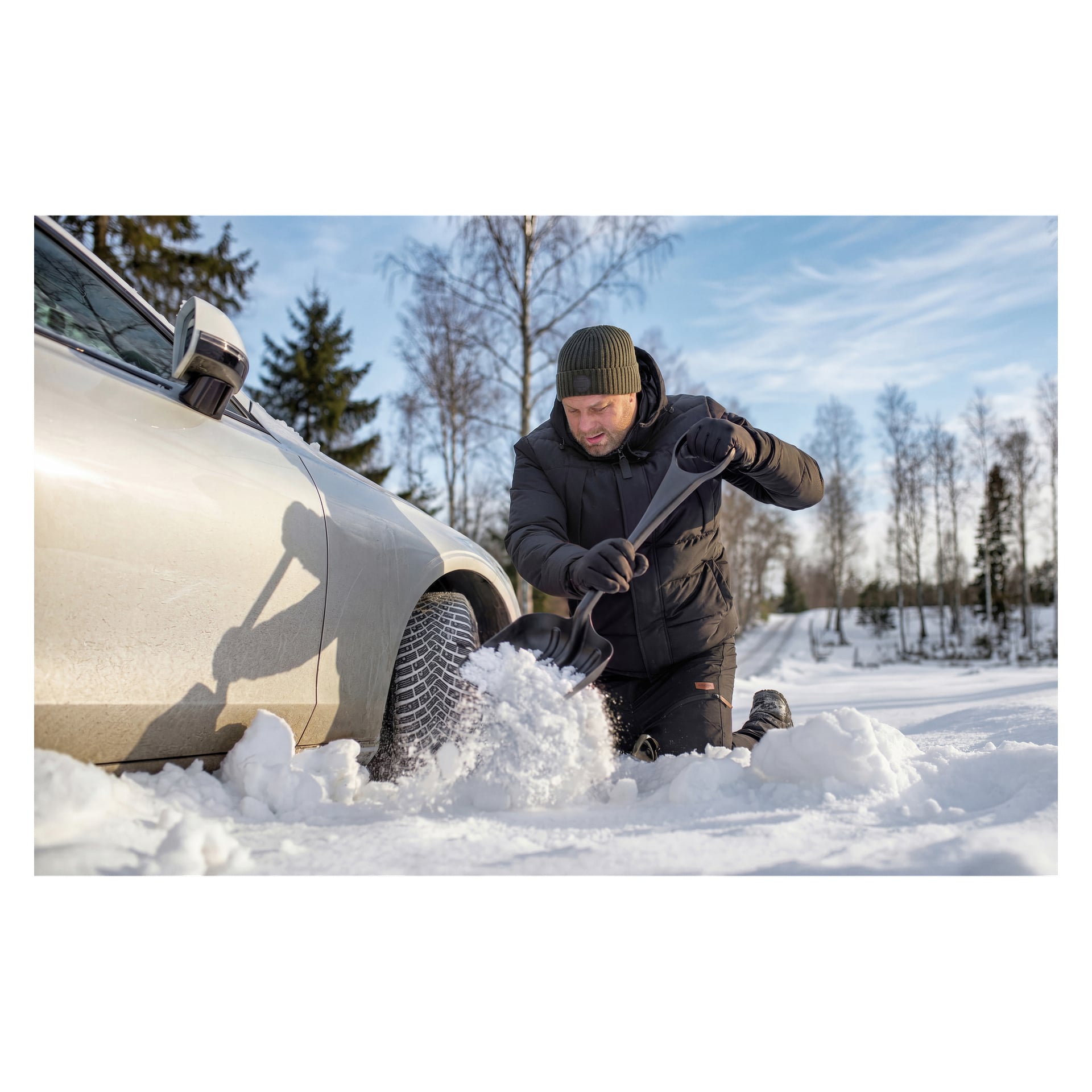 En person i vinterkläder knäböjer bredvid en bil en solig, snöig dag och röjer snö från ett däck med en Hamron snöspade. I bakgrunden syns snötäckta träd och en blå himmel.
