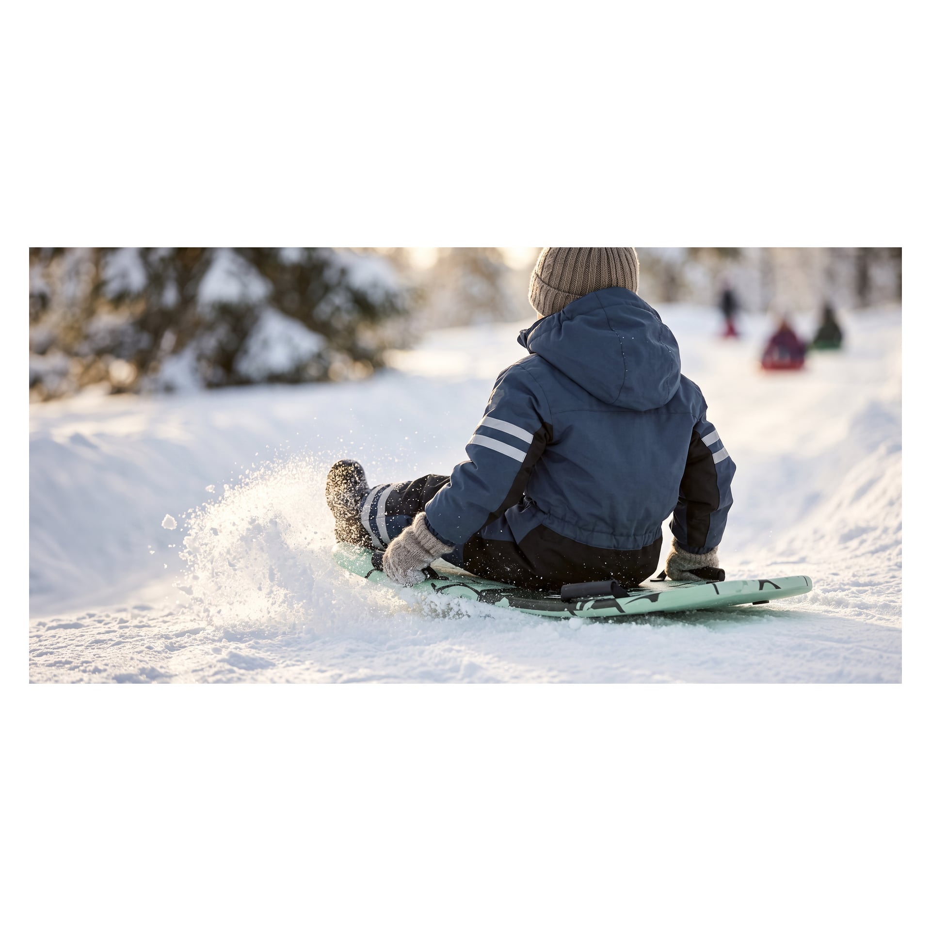 Ein Kind in Winterjacke und Mütze rutscht auf einem hellgrünen Plastik-Kayoba-Snowboard einen verschneiten Hügel hinunter.