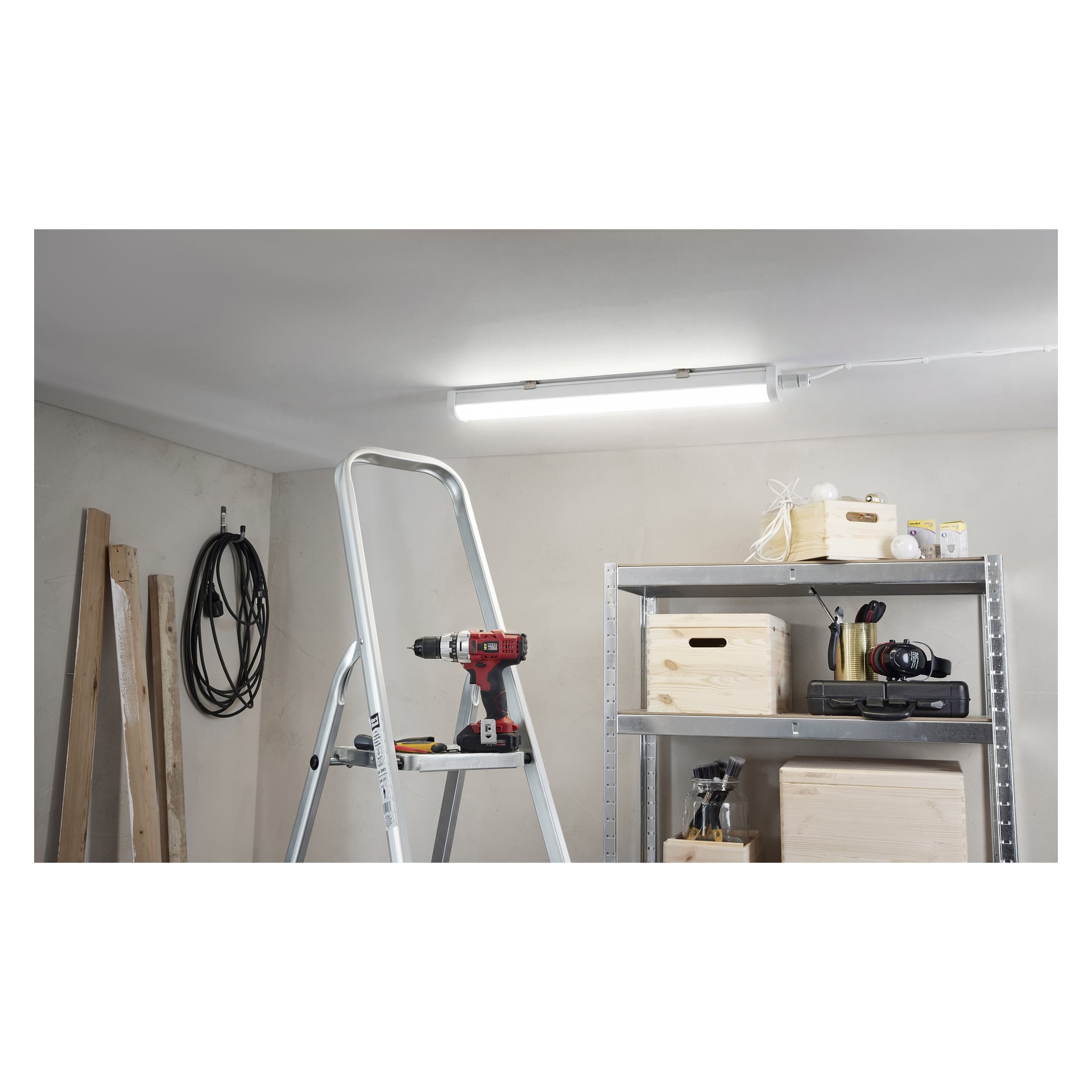 Eine Trittleiter steht in einer Garage neben einer Bohrmaschine, Metallregale mit Kisten, zusätzliche Glühbirnen, eine Kamera, aufgewickelte Kabel an der Wand und Holzplanken, die in der Ecke unter einer "Anslut LED-Lampe" stehen.