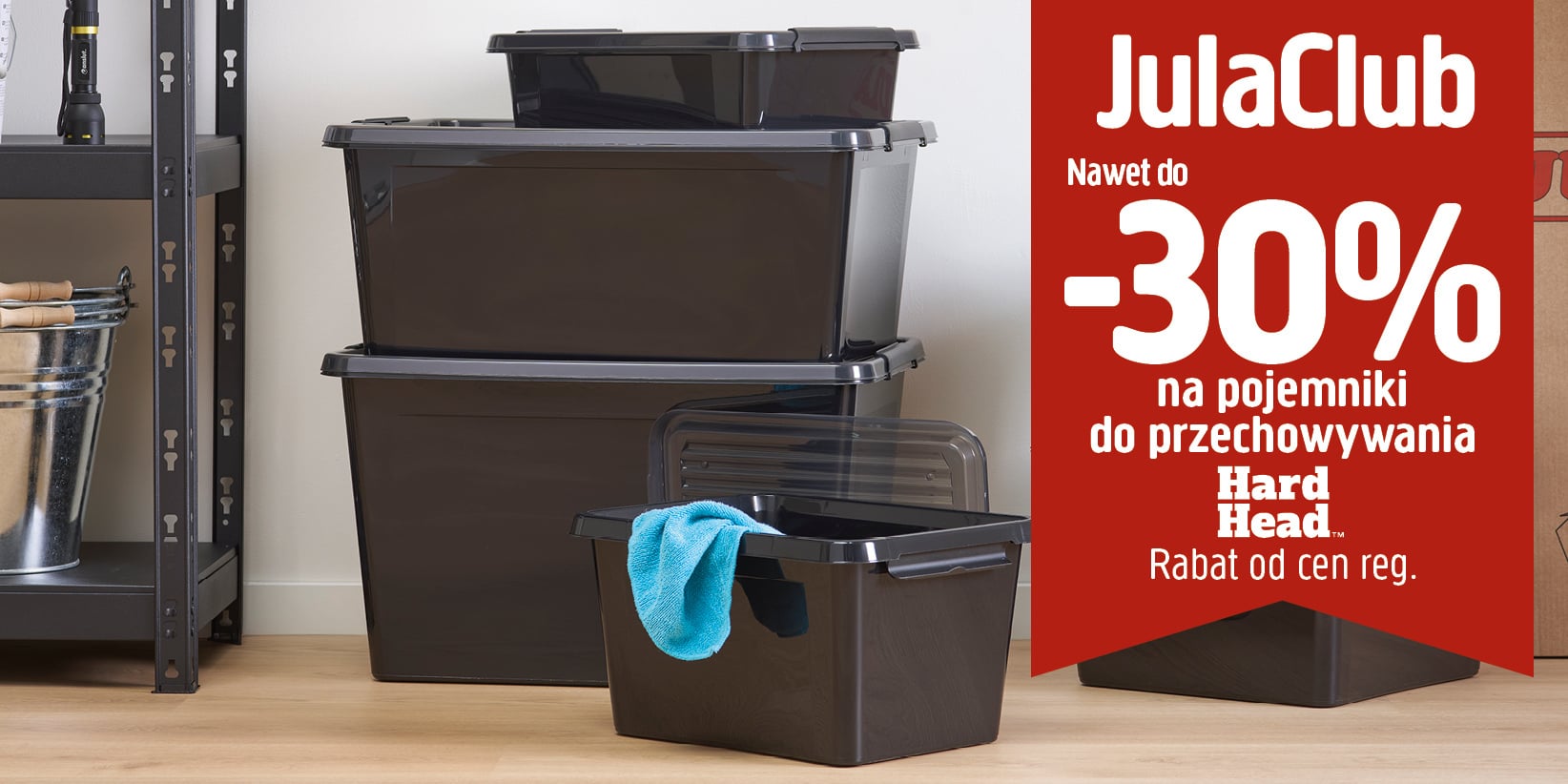 Trzy ułożone jedna na drugiej czarne plastikowe pojemniki do przechowywania obok czerwonego plakatu reklamowego z napisem: JulaClub Nawet do -30% na pojemniki do przechowywania Hard Head. Rabat od cen regulaminowych.