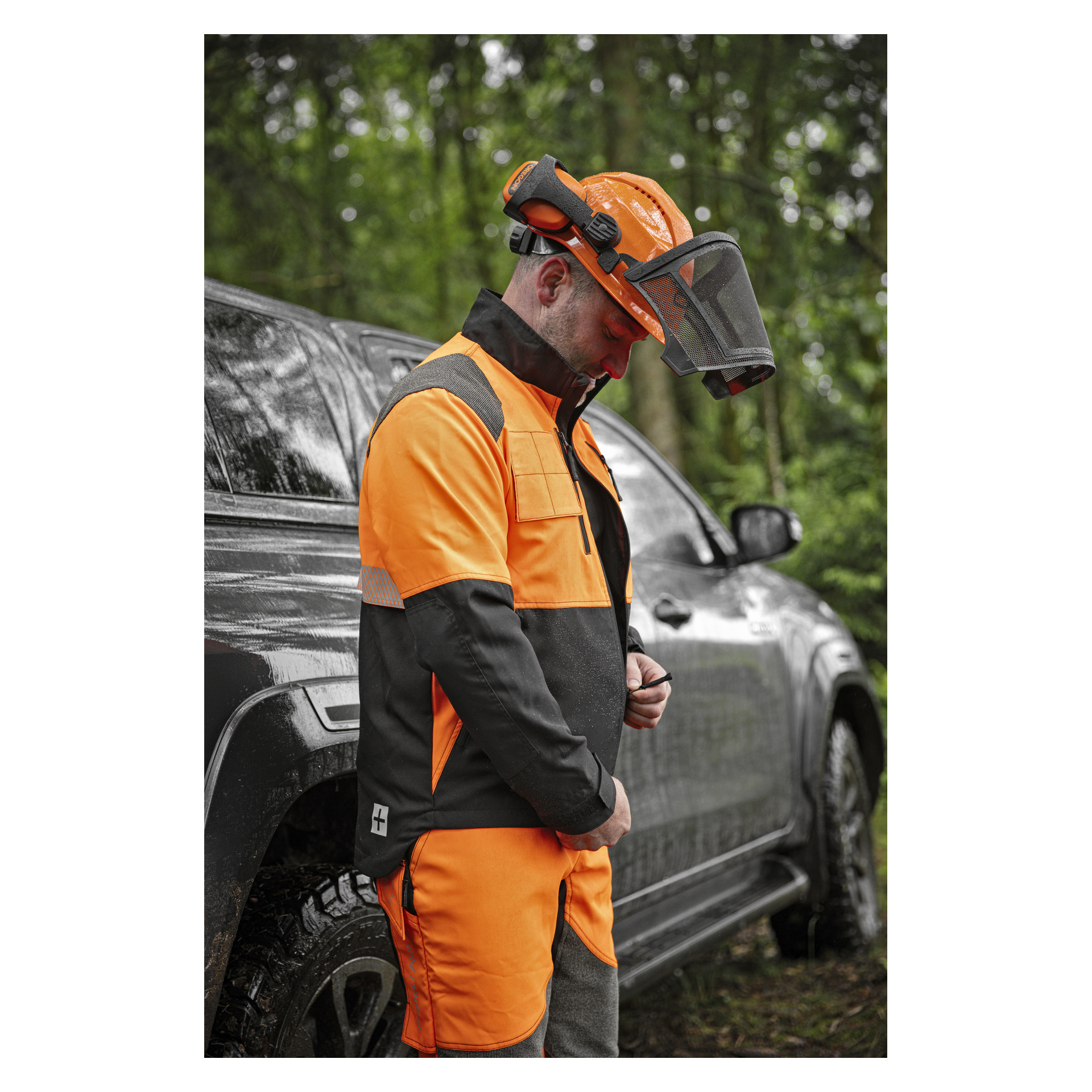 En person som bär en Protect Plus högsynlig orange och svart skyddsjacka samt matchande byxor, tillsammans med hjälm med ansiktsskydd och hörselkåpor, står bredvid ett fordon.