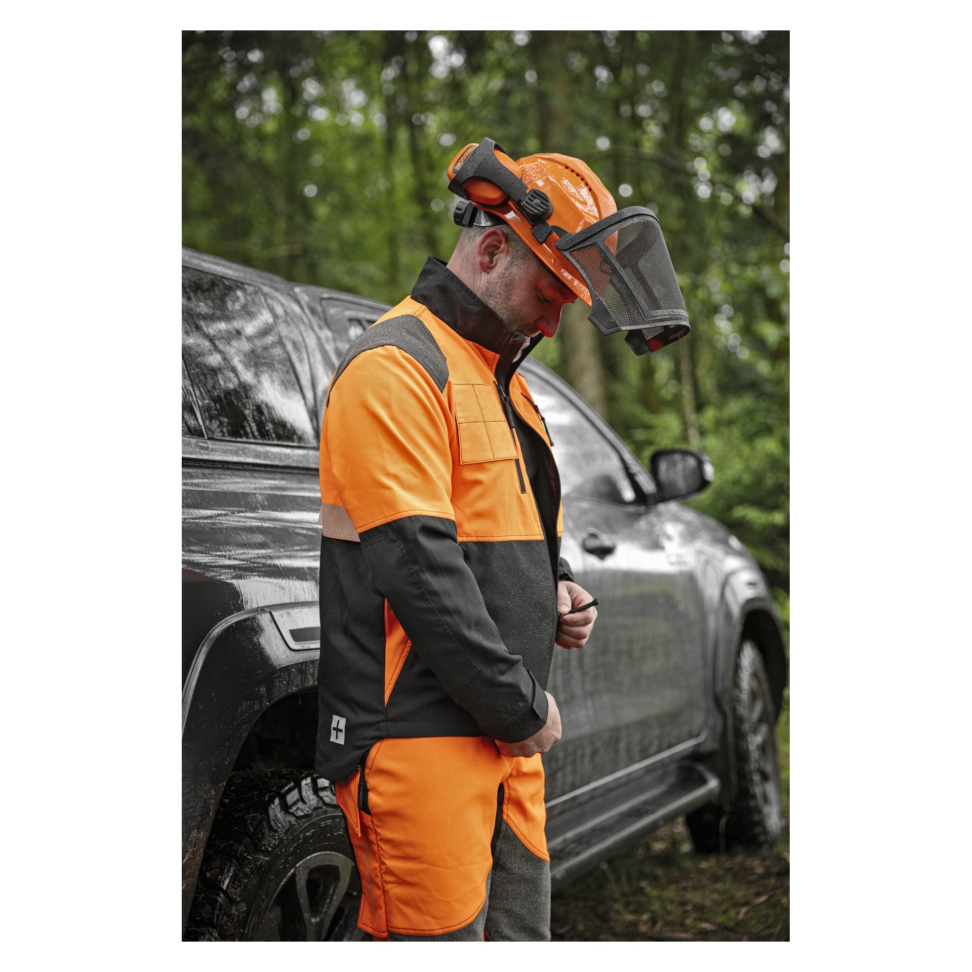 En arbetare som bär en Oregon Protect Plus högsynlig orange och svart skyddsjacka, byxor och hjälm med ansiktsskydd, står bredvid ett fordon.