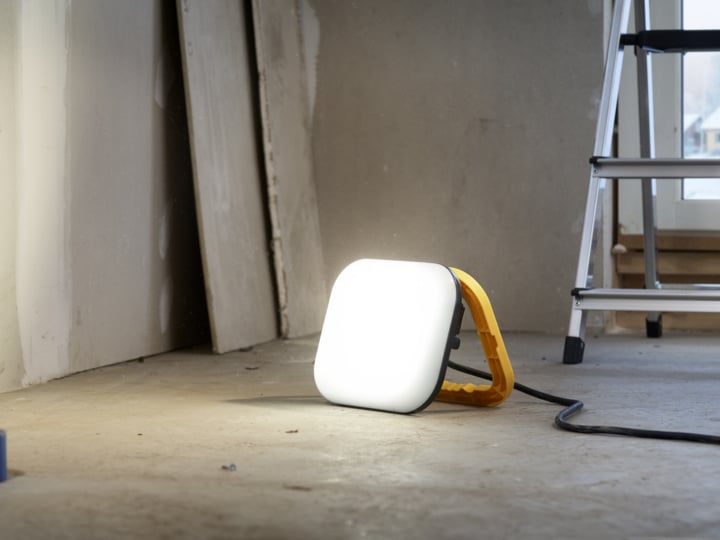 Eine tragbare LED-Arbeitslampe mit gelbem Griff steht auf einem Betonboden neben einer Trittleiter.