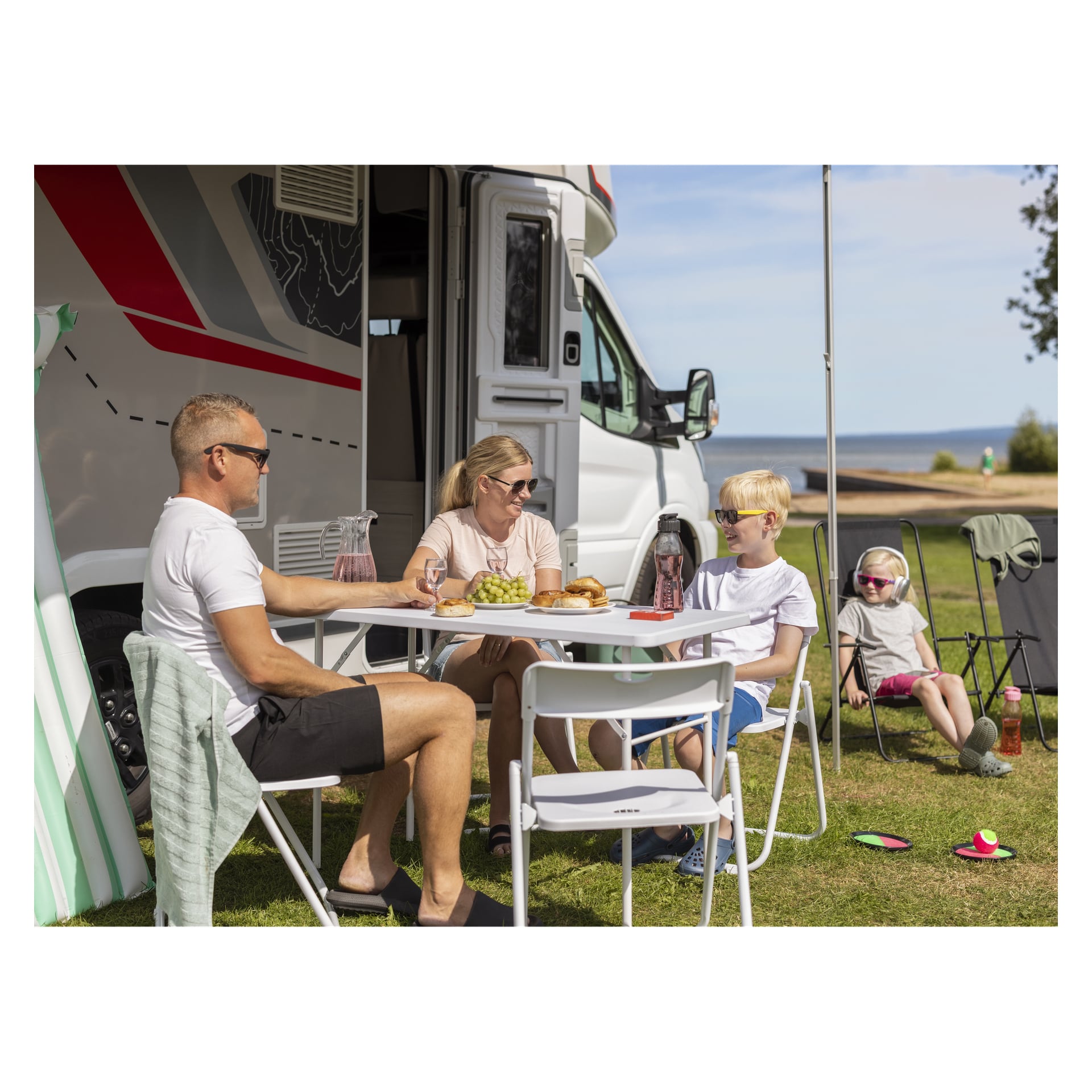 Eine Familie sitzt an einem Klapptisch vor einem Freizeitfahrzeug, während ein Mädchen mit Kopfhörern in einem EKVIP-Klappstuhl in der Nähe auf dem Gras sitzt.