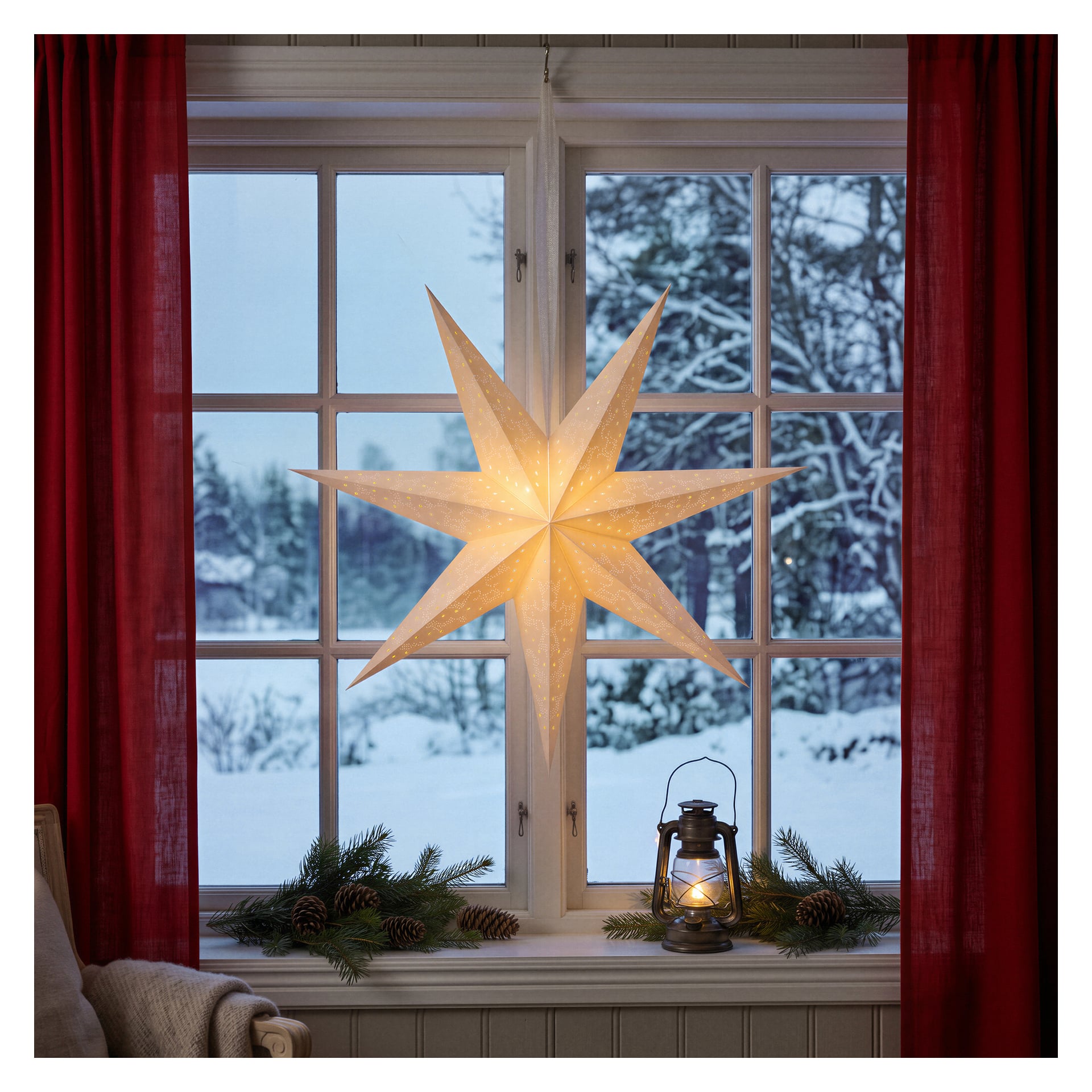En upplyst pappersstjärndekoration, upplyst av en EKVIP Rechargeable LED-lampa, hänger i ett fönster framför ett snötäckt landskap, med en liten lykta och tallgrenar på fönsterbrädan.