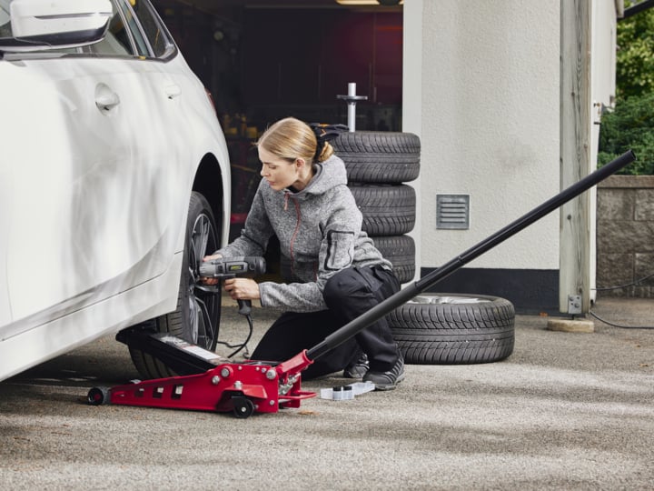 Eine Frau benutzt ein Elektrowerkzeug, um Radmuttern von einem Autorad zu entfernen, während das Fahrzeug von einem roten hydraulischen Wagenheber getragen wird.