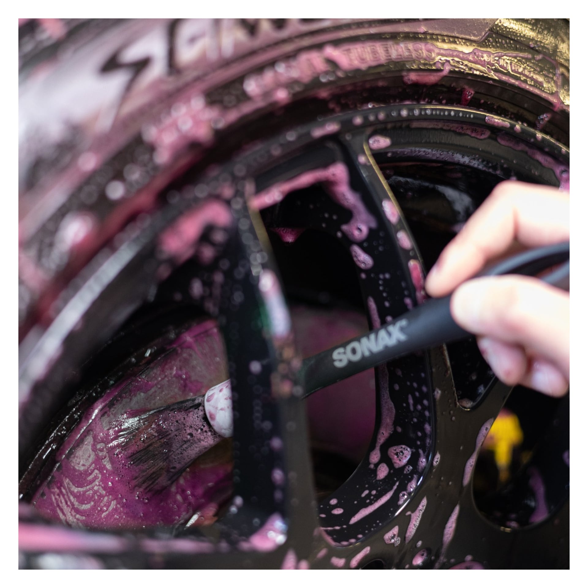 En hand använder en Sonax Deep cleaning brushes Profiline 2-pack för att rengöra ett svart bilhjul täckt med rosa rengöringsskum.