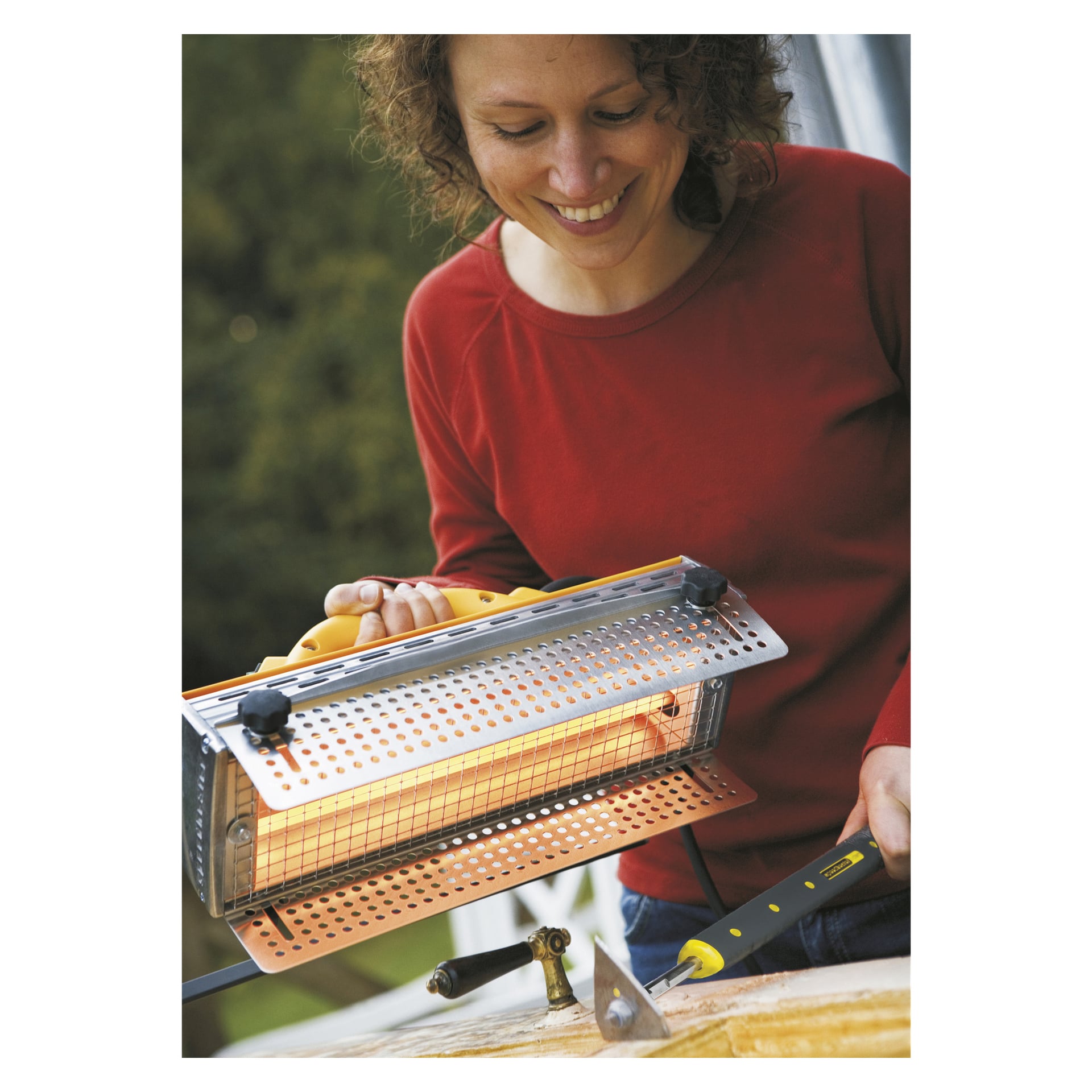 En smilende person i rød skjorte holder Speedheater-standarden over en treflate, som lyser oransje for å indikere at den er slått på, mens den holder en skraper med den andre hånden.