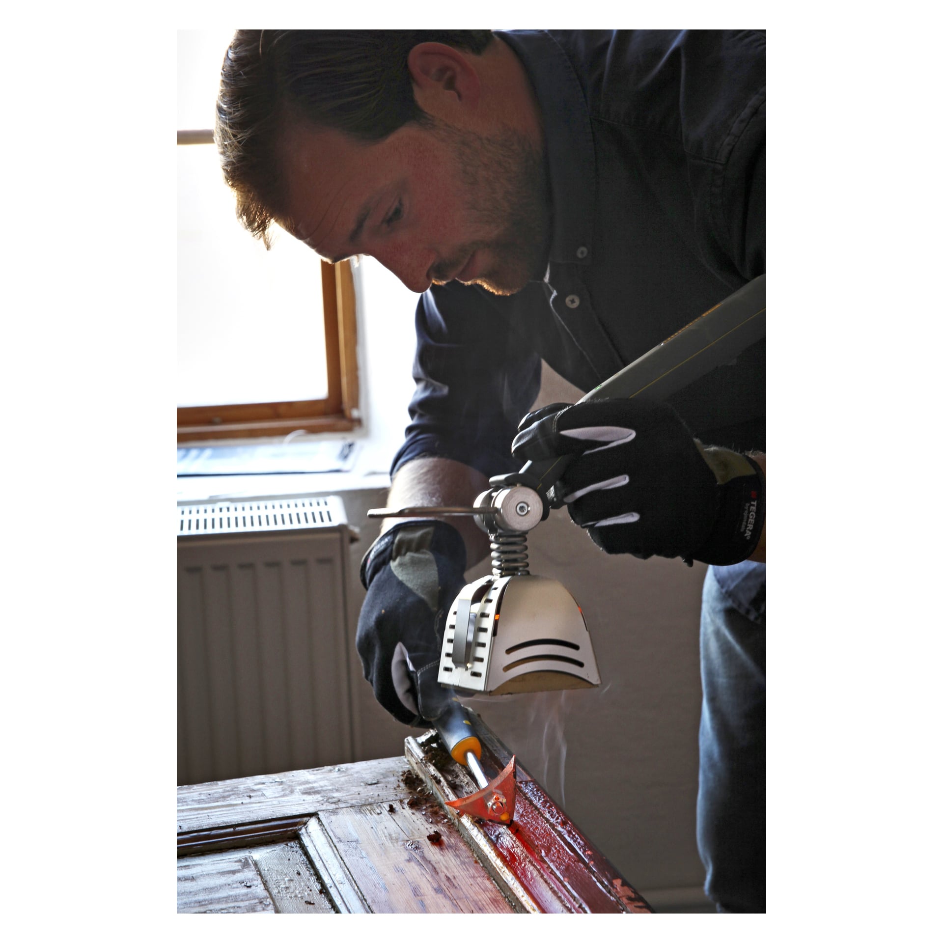 Hanskat päässä oleva henkilö poistaa maalia puuovesta ikkunan lähellä käyttäen Speedheater Cobraa ja raapimistyökalua.