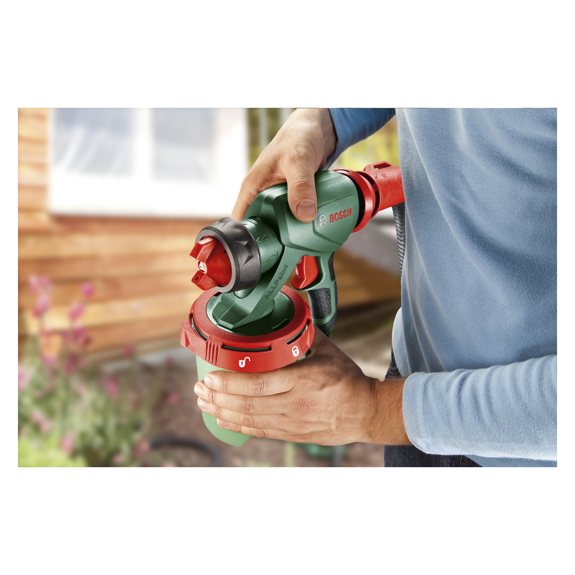Noen i blå skjorte kobler den røde og grønne dysen på en Bosch spraypistol PFS 5000 E til beholderen, mens et uklart hus og en hage dukker opp bak dem.