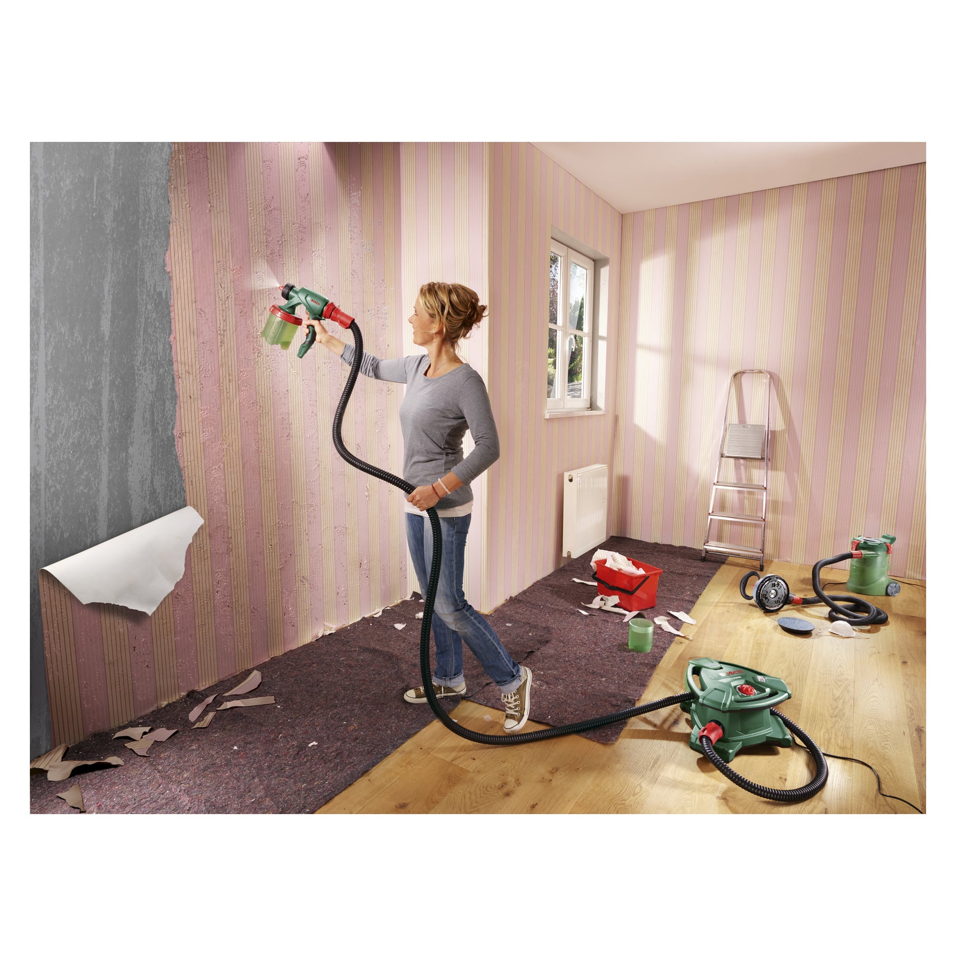 En kvinne bruker en Bosch spraypistol PFS 5000 E mens hun fjerner rosa stripete tapet fra en vegg i et godt opplyst rom. Biter av revet tapet og ulike verktøy ligger spredt utover gulvet, med en stige og ekstra forsyninger i bakgrunnen.