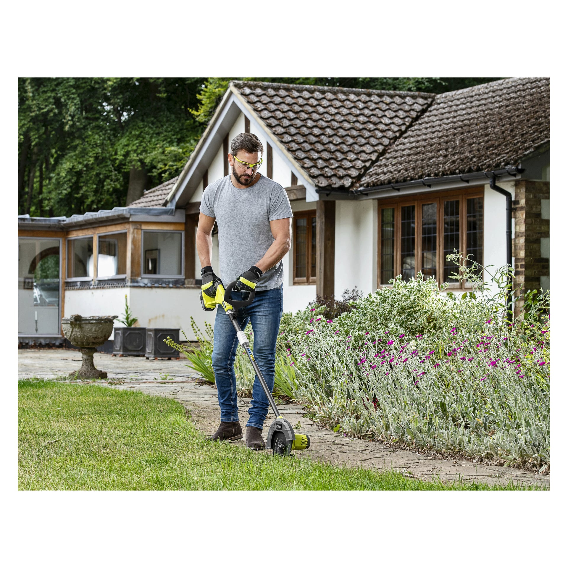 En mann kledd i vernebriller, hansker, jeans og grå skjorte bruker en Ryobi One+ Edge-trimmer RY18EGA-0 for å trimme kanten av en plen ved siden av en blomsterbed utenfor et hus med tegltak og store vinduer.