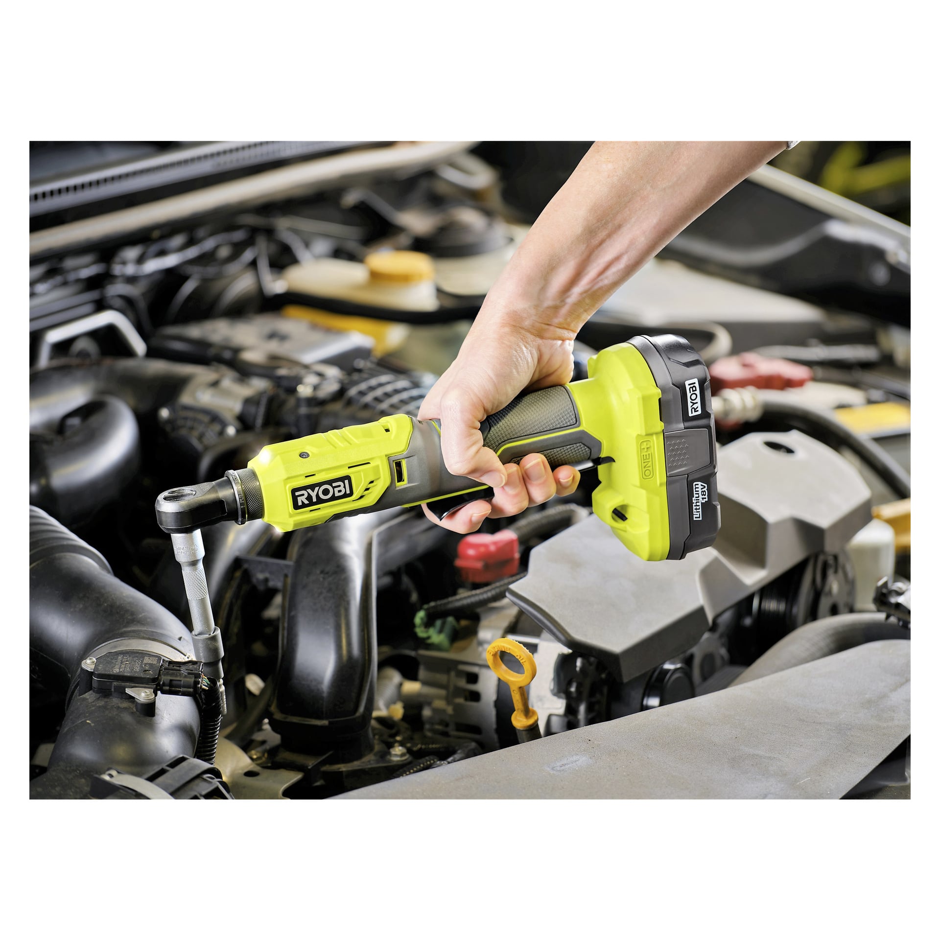 En person betjener et grønt Ryobi One+ batteridrevet skrallehåndtak R18RW2-0 for å jobbe på en bolt under det åpne panseret på en bil, med ulike motordeler og oljepeilepinnen synlig i scenen.