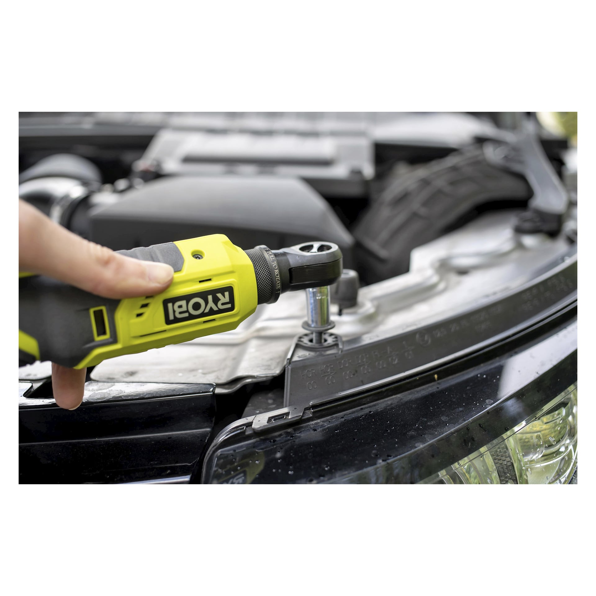 Noen bruker et gult Ryobi One+ batteridrevet skrallehåndtak (modell R18RW2-0) med en pipe festet, og strammer en bolt i motorrommet på en bil. Bilens panser er åpent, og viser ulike motorkomponenter.