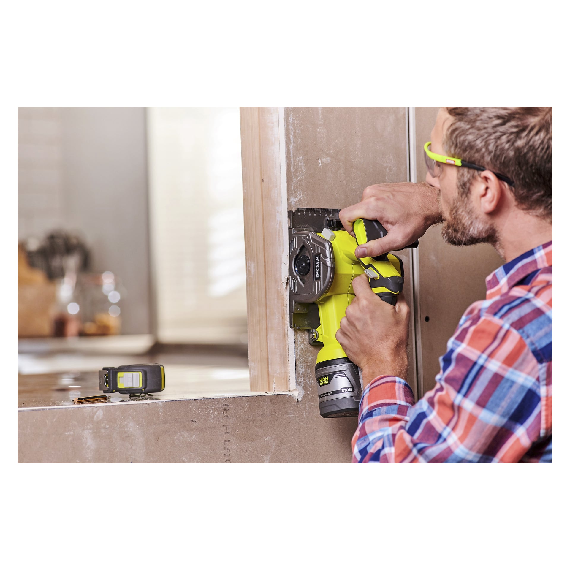 En person med skyddsglasögon skär gipsskivor nära en dörrkarm med det gröna Ryobi One+ Multi-saw R18MMS-0 sladdlösa elverktyget. En kompakt lasermätare står på den närliggande fönsterkarmen.