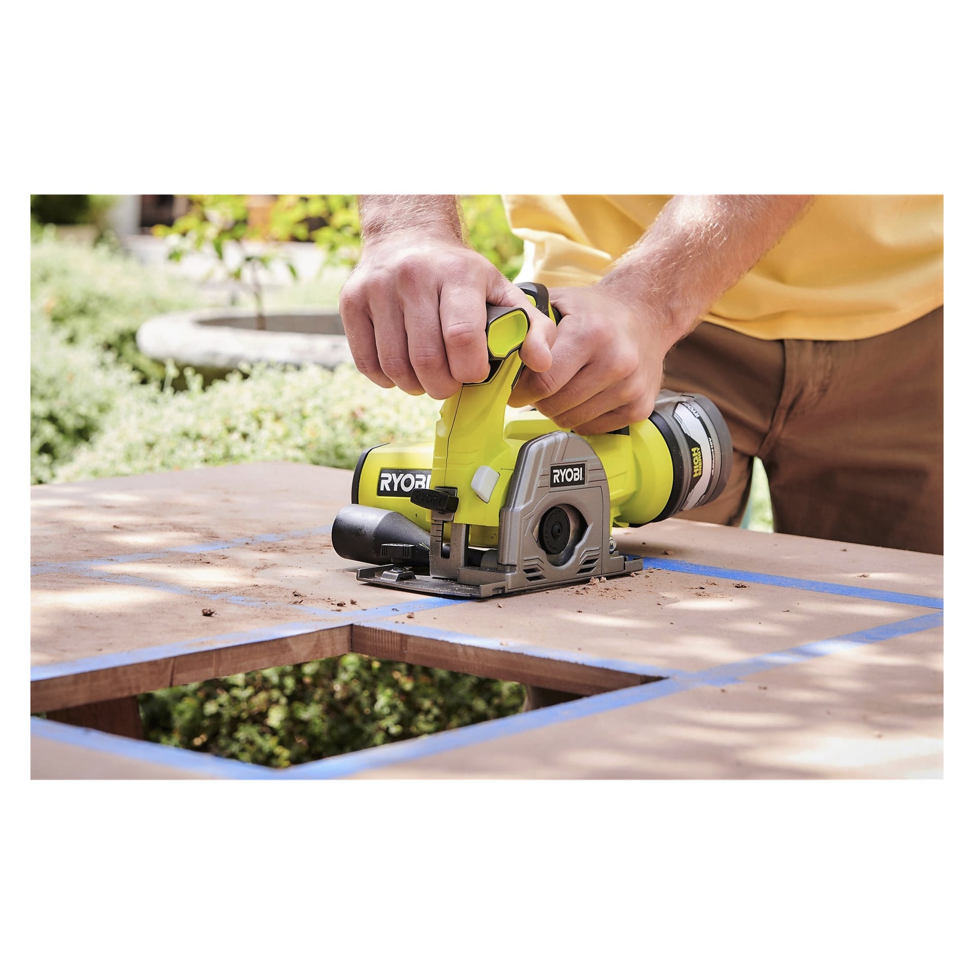 Någon använder en Ryobi One+ Multi-saw R18MMS-0 sladdlös minicirkelsåg för att kapa en rektangulär form i en träskiva fodrad med blå tejp, utomhus bland grön vegetation.