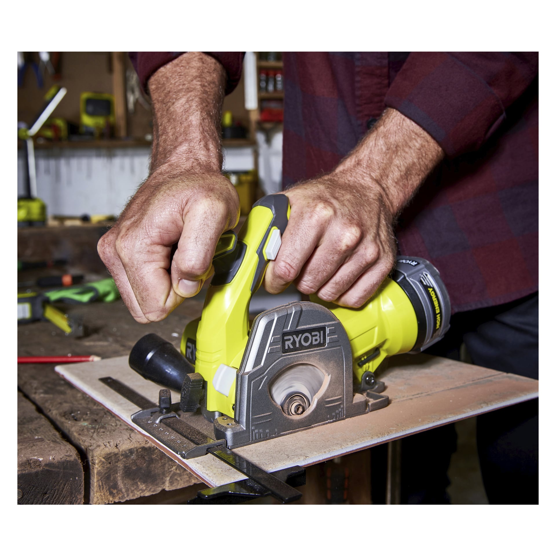 En person håller i en gul Ryobi One+ Multi-saw R18MMS-0 med båda händerna och sågar trä i en verkstad med olika verktyg som syns på arbetsbänken bakom dem.