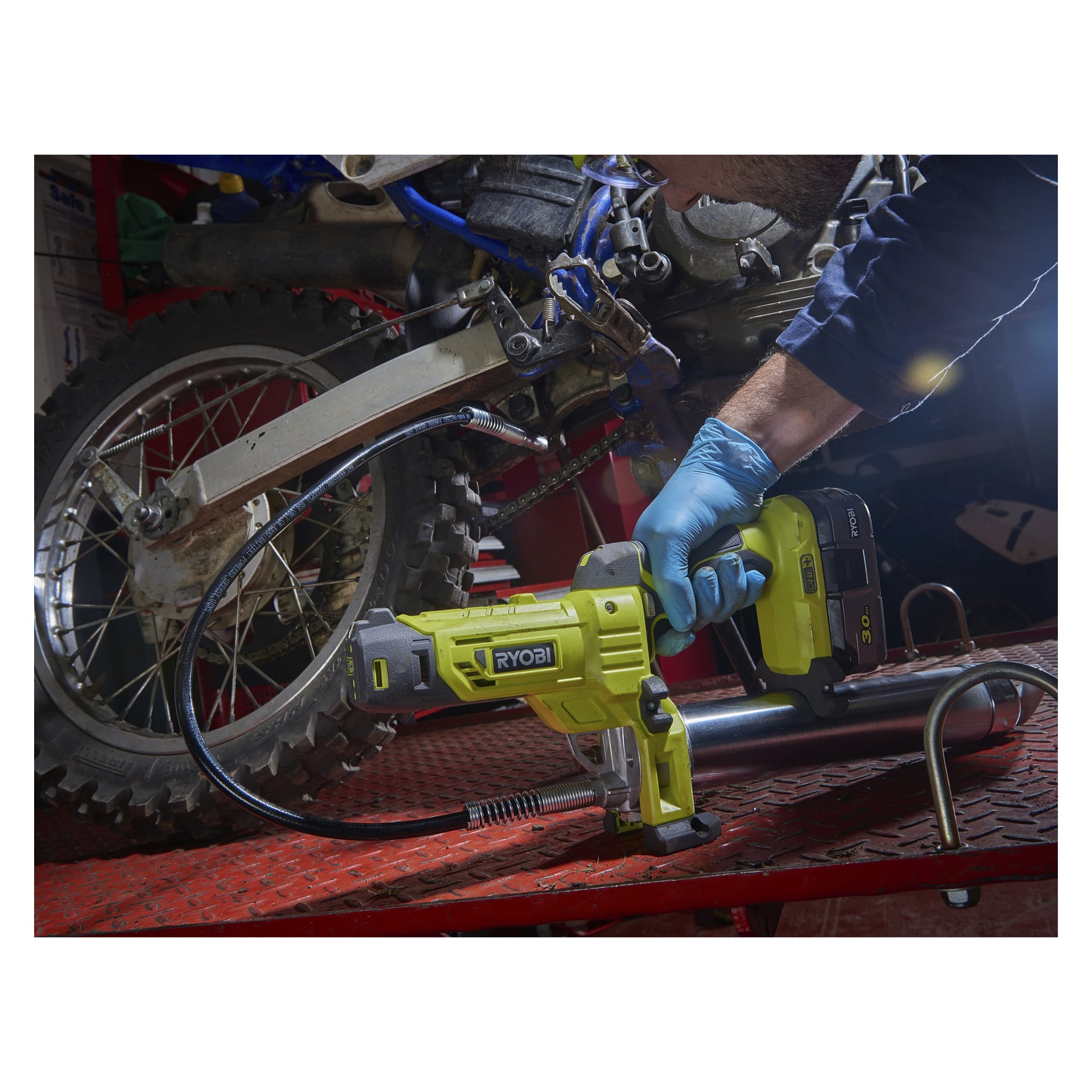 Med blå hansker bruker man en Ryobi One+ Grease Gun R18GG-0 for å vedlikeholde bakhjulet på en crossmotorsykkel. Aktiviteten foregår på en rød plattform inne i et sterkt opplyst verksted, hvor ulike verktøy er synlige i bakgrunnen.