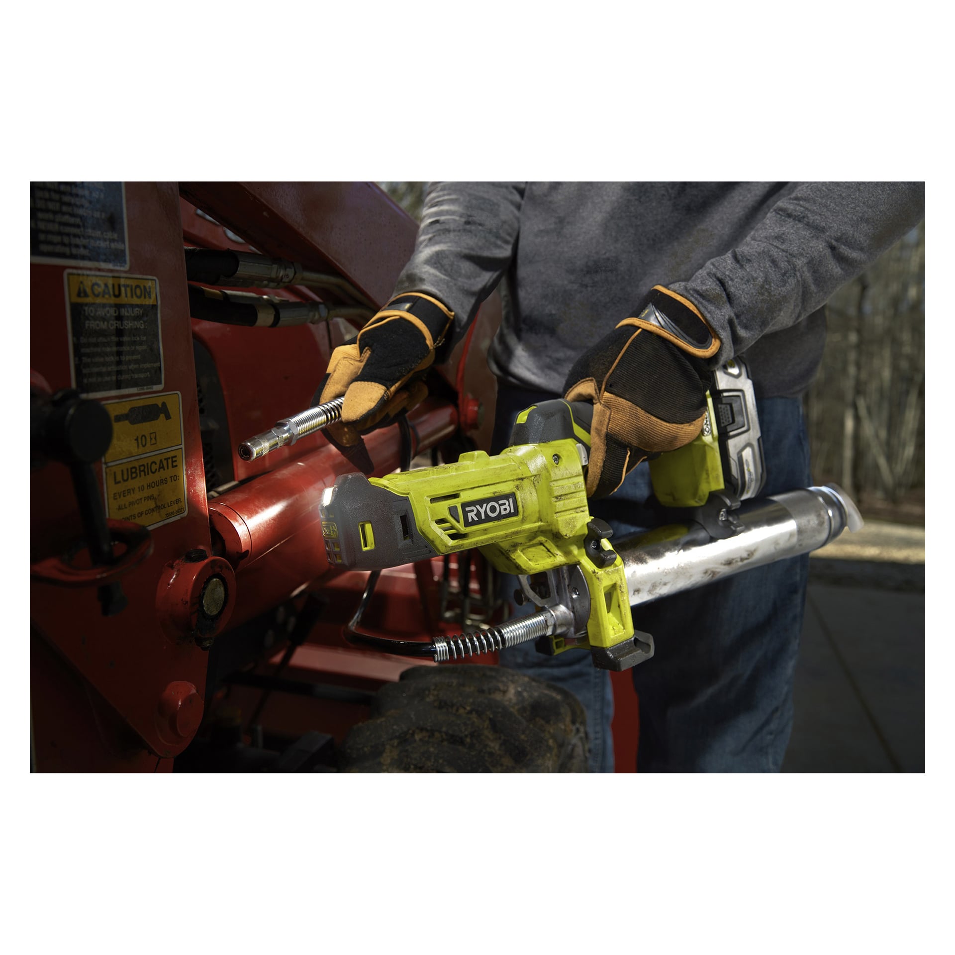 En person med hansker betjener en Ryobi One+ fettpistol R18GG-0 for å påføre fett på maskinens metallskjøt, og sikter dysen nær en synlig advarselsetikett.