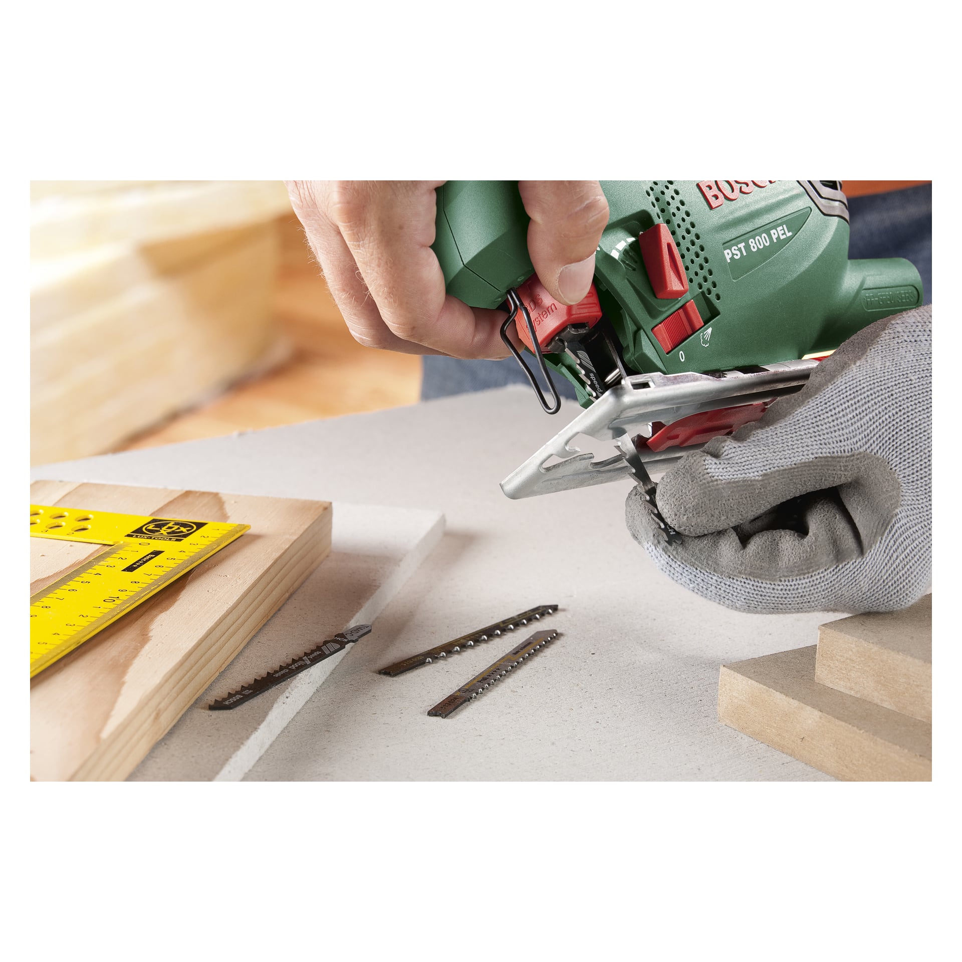 Rękawiczka chwyta zieloną wyrzynarkę Bosch PST 800 PEL, gotową do włożenia ostrza. W pobliżu na powierzchni roboczej spoczywają trzy dodatkowe ostrza wyrzynarki i żółta linijka kątna, obok kilku drewnianych elementów.
