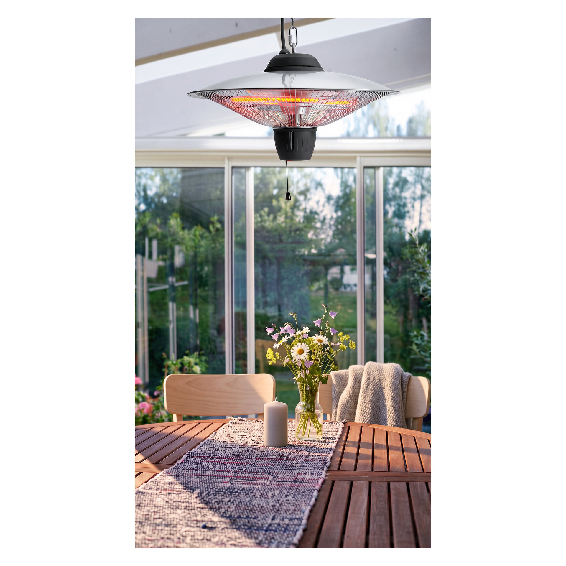 Pyöreä terassipöytä on koristeltu teksturoidulla juoksijalla, sytetyllä kynttilällä ja maljakolla täynnä villikukkia, jotka on sijoitettu Anslut Hanging Infrared Heaterin alle aurinkohuoneeseen. Suuret ikkunat ympäröivät tilaa ja tarjoavat näkymiä rehevälle vehreydelle ulkona.