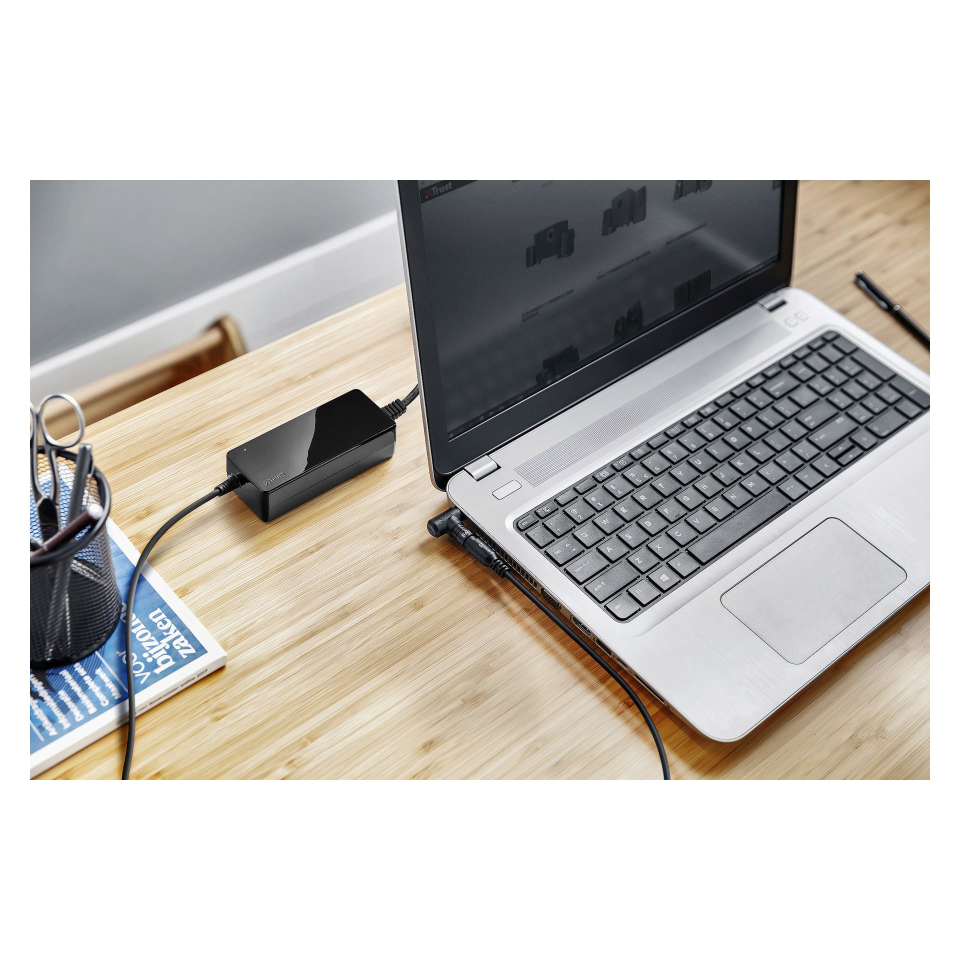 Na drewnianym biurku leży otwarty srebrny laptop, podłączony do czarnej ładowarki Trust. Obok niego stoi uchwyt na długopisy wypełniony długopisami oraz magazyn. Na ekranie wyświetlana jest strona internetowa w odcieniach szarości.